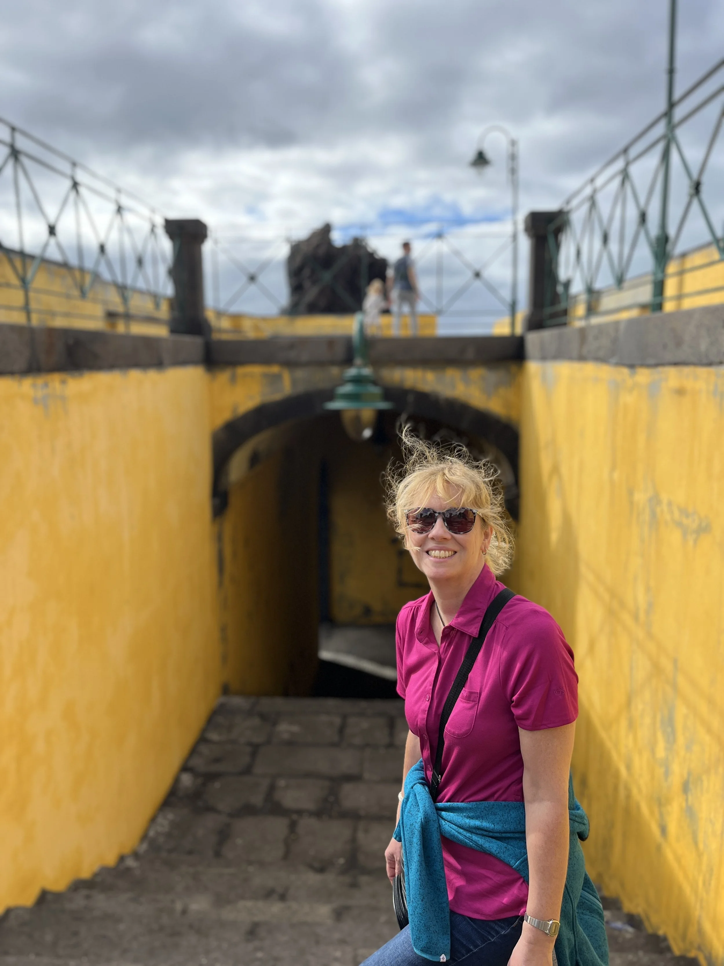 A woman with blonde hair, wearing sunglasses and a bright pink shirt, smiling at the camera, standing in front of a yellow wall with an archway, with a person and a child visible in the background on a bridge under a cloudy sky.
