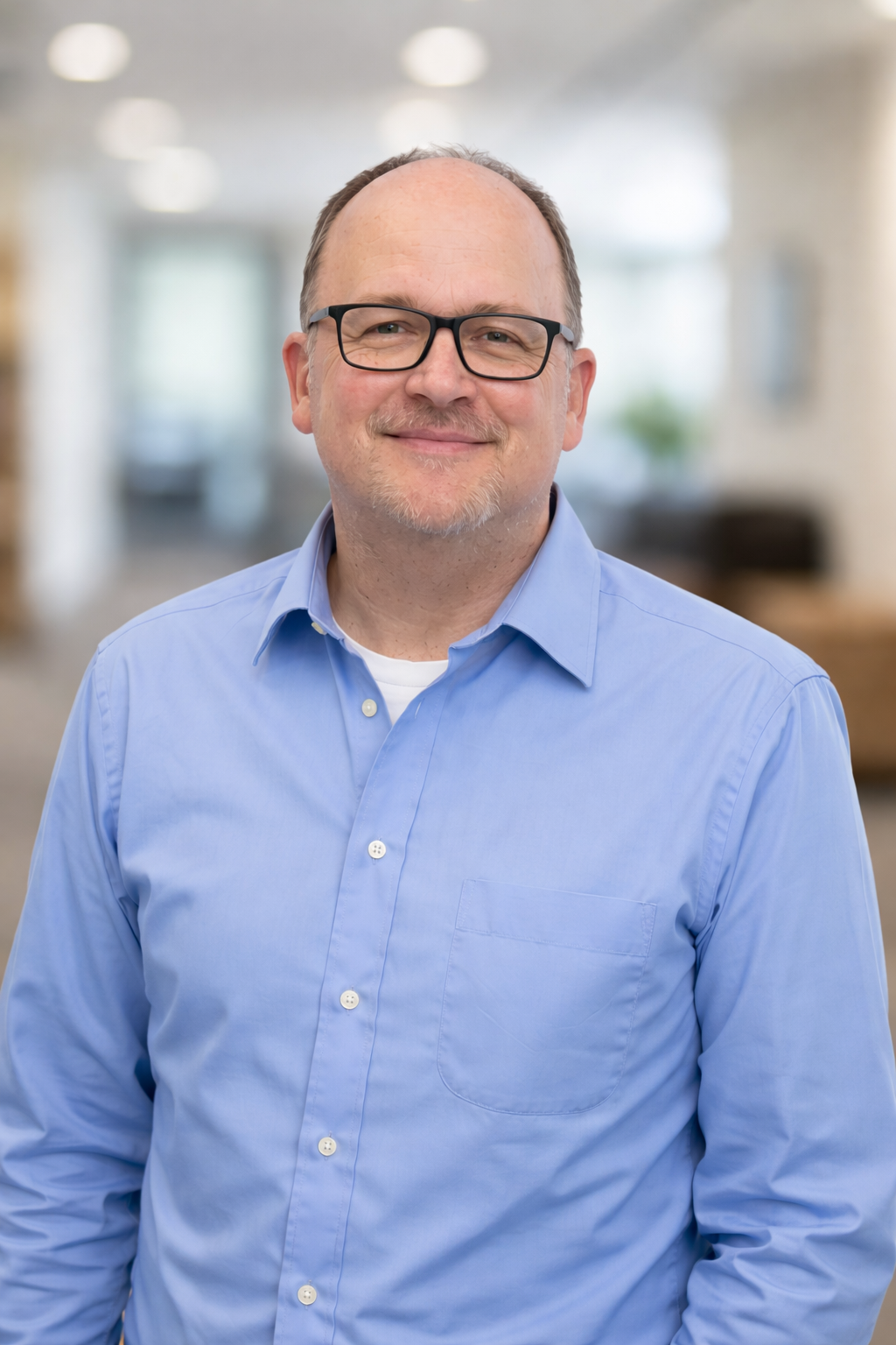 A middle-aged man with glasses and a light beard, wearing a blue button-down shirt, smiling in a professional indoor setting.