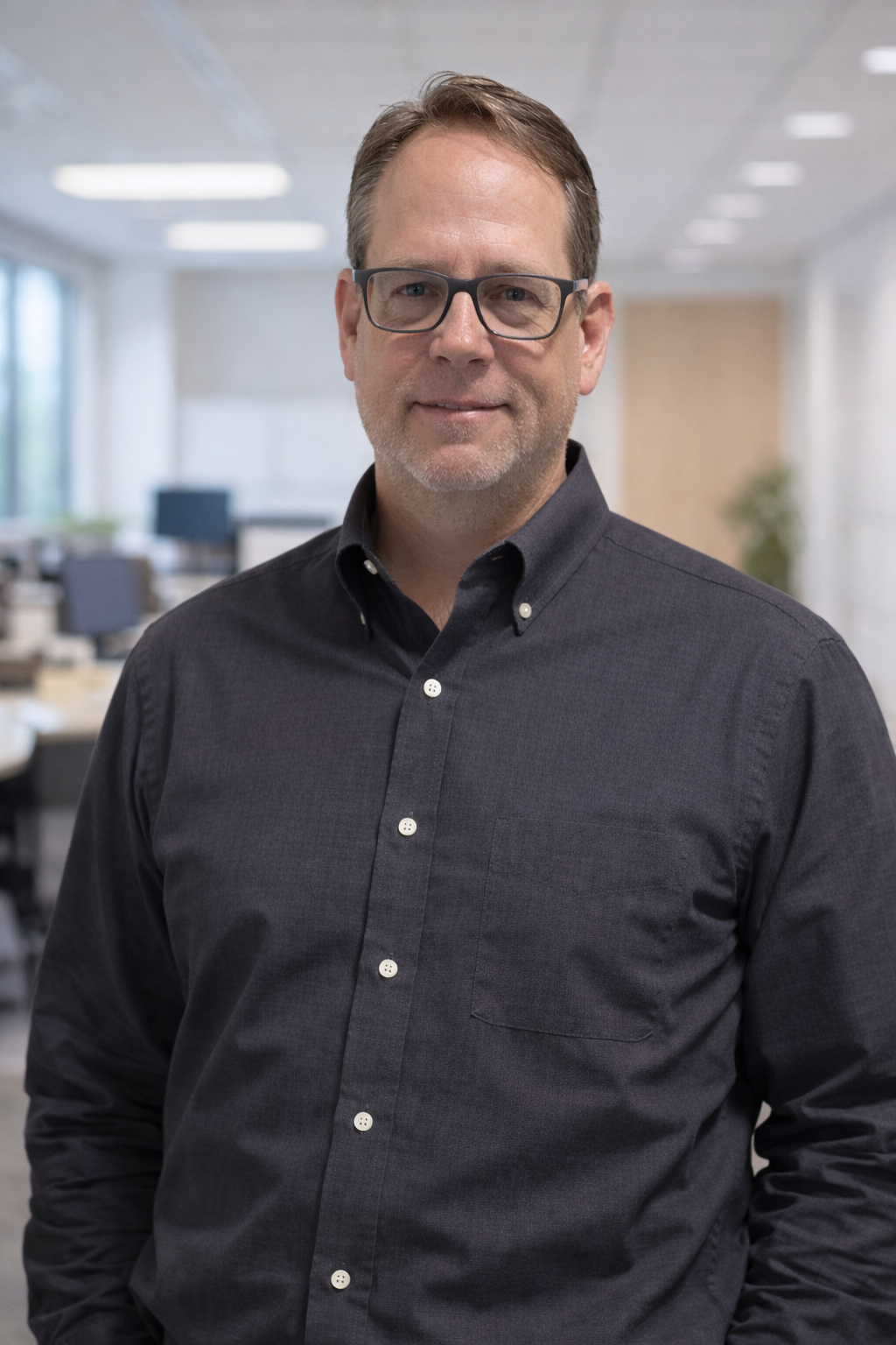 A man with glasses and a beard wearing a dark button-up shirt standing in an office setting.