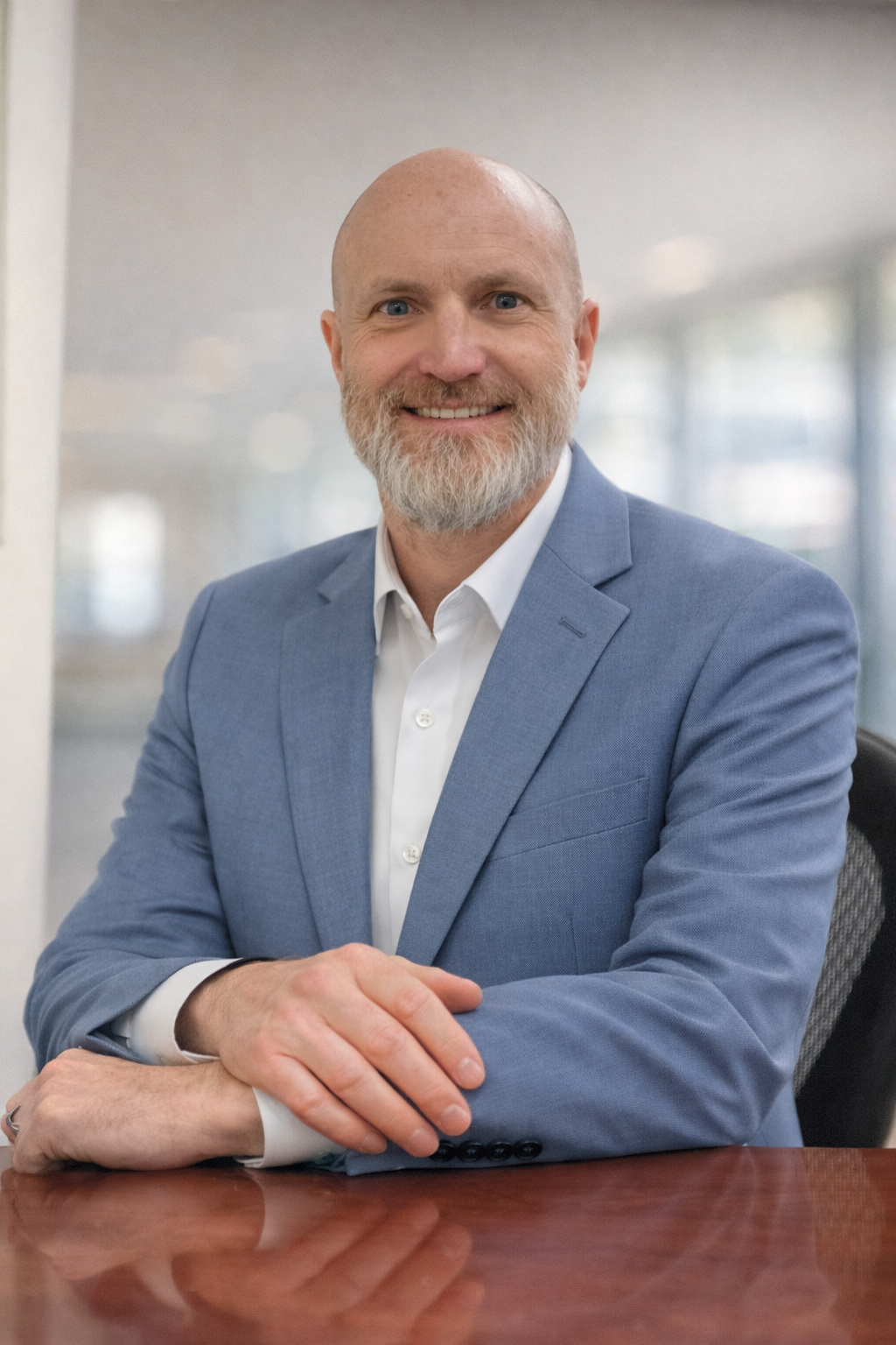 A middle-aged man with a beard and a shaved head, wearing a light blue suit and a white shirt, sitting at a wooden conference table in an office setting, smiling at the camera.