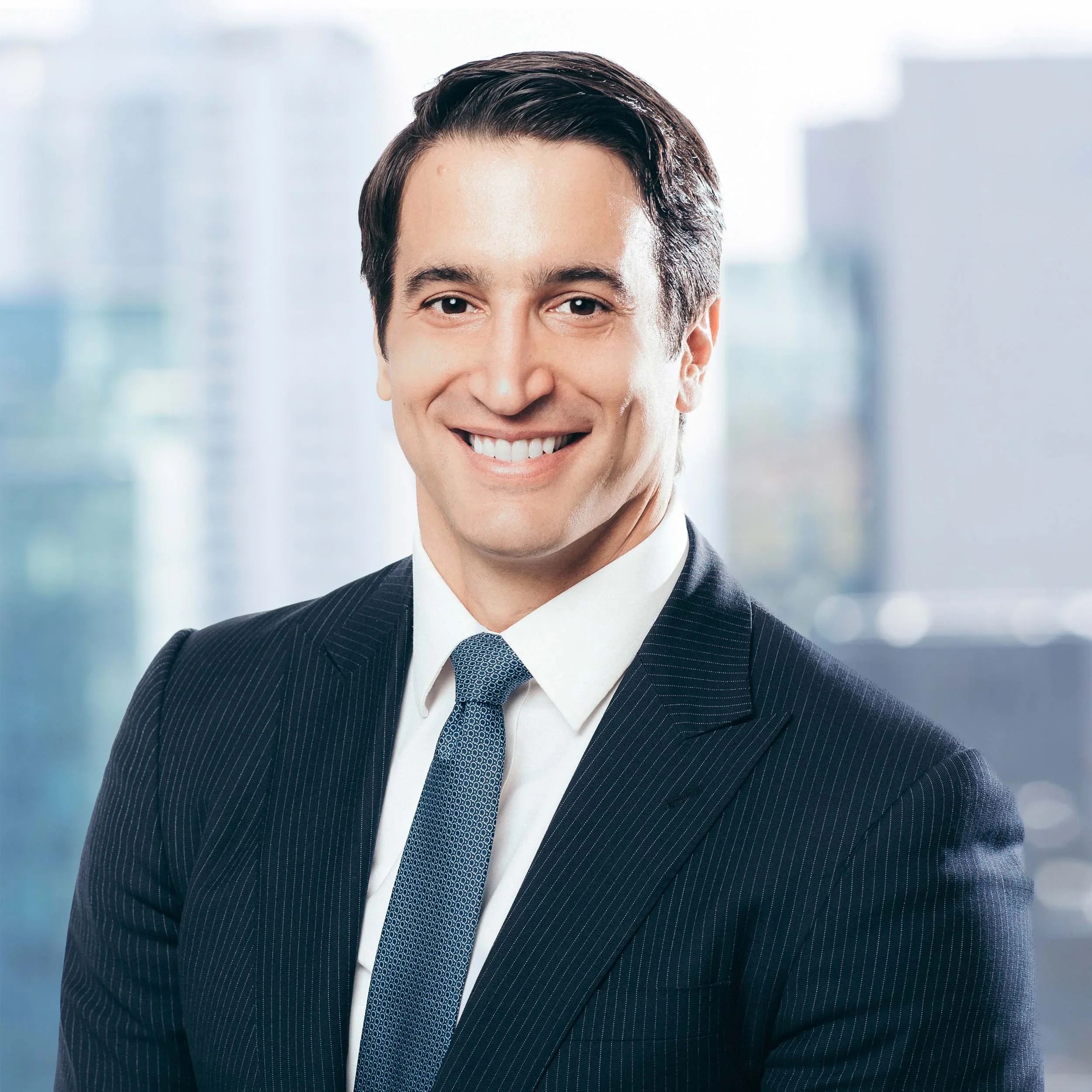 A young man with dark hair, wearing a dark suit and tie, smiling in front of a cityscape background.