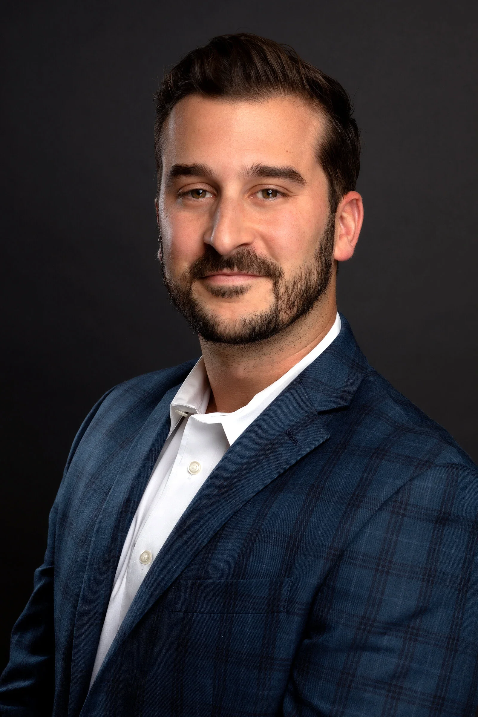 A professional headshot of a man with brown hair, a beard, wearing a dark blue plaid blazer and white shirt, against a dark background.