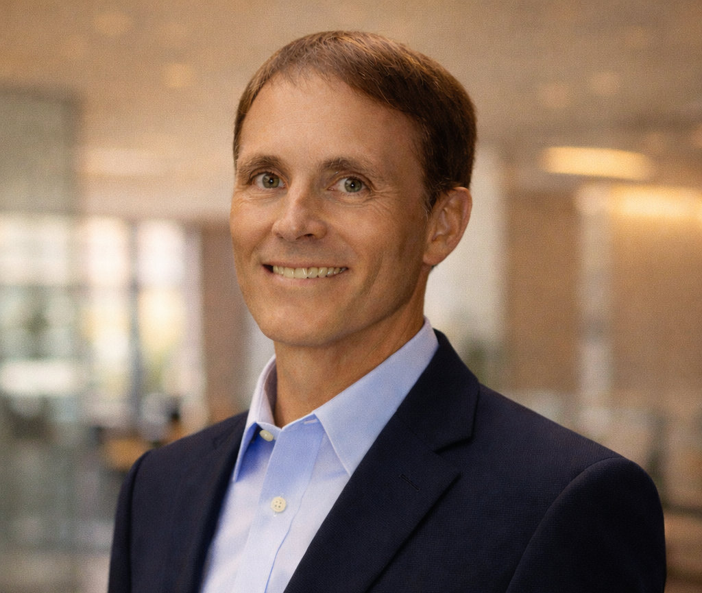 A professional man in a dark suit and light blue shirt smiling indoors.