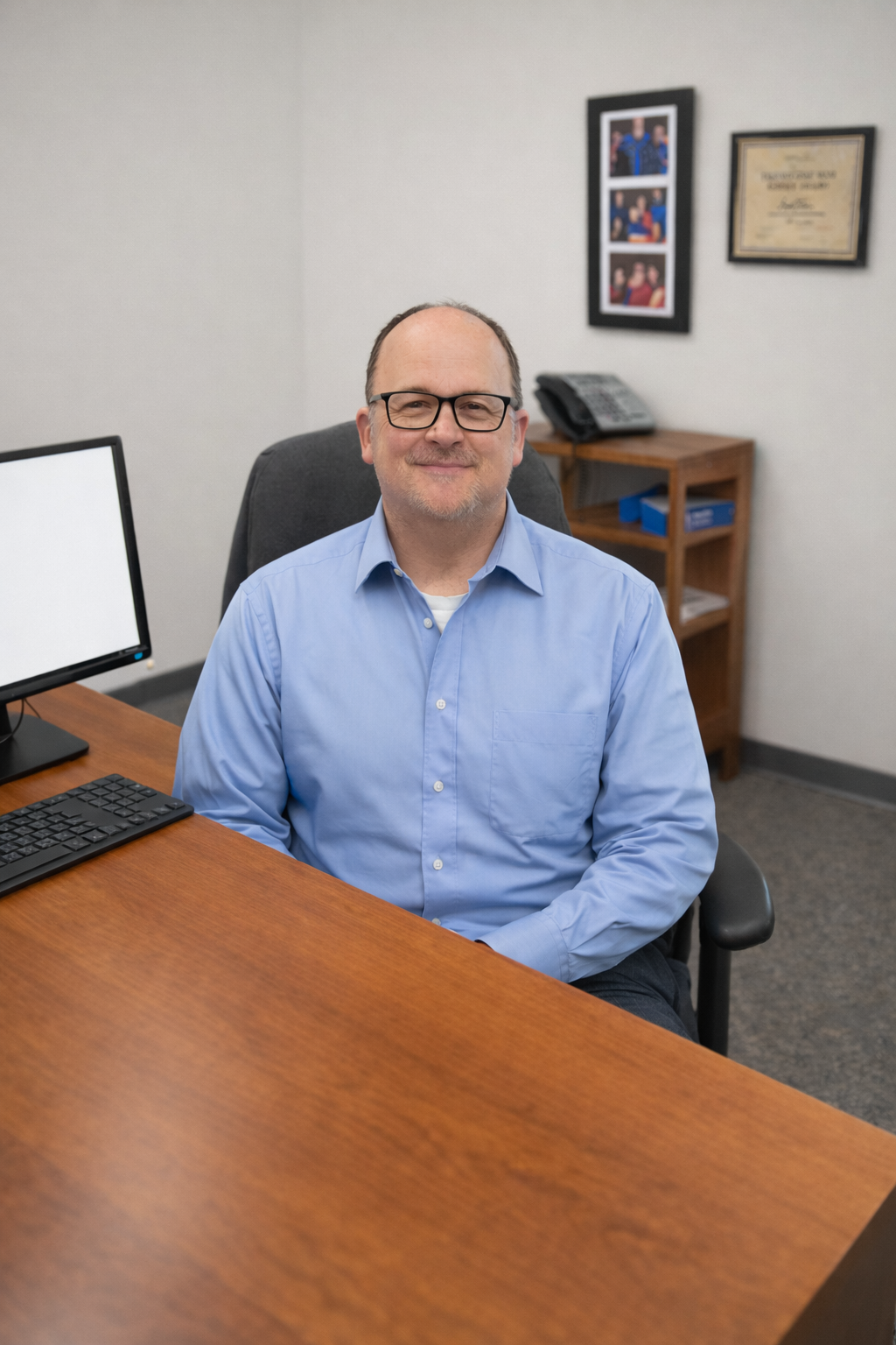 A man with glasses and a blue shirt sitting at a desk in an office, smiling.