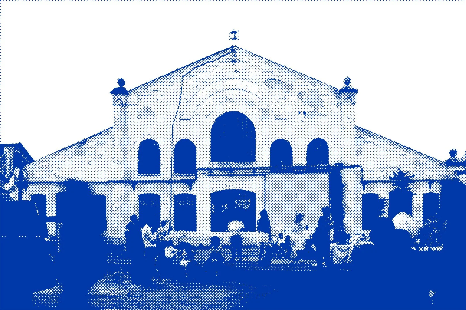 A large building with an arched entrance and multiple windows, with decorative figures on the roof, and people standing and sitting in front.