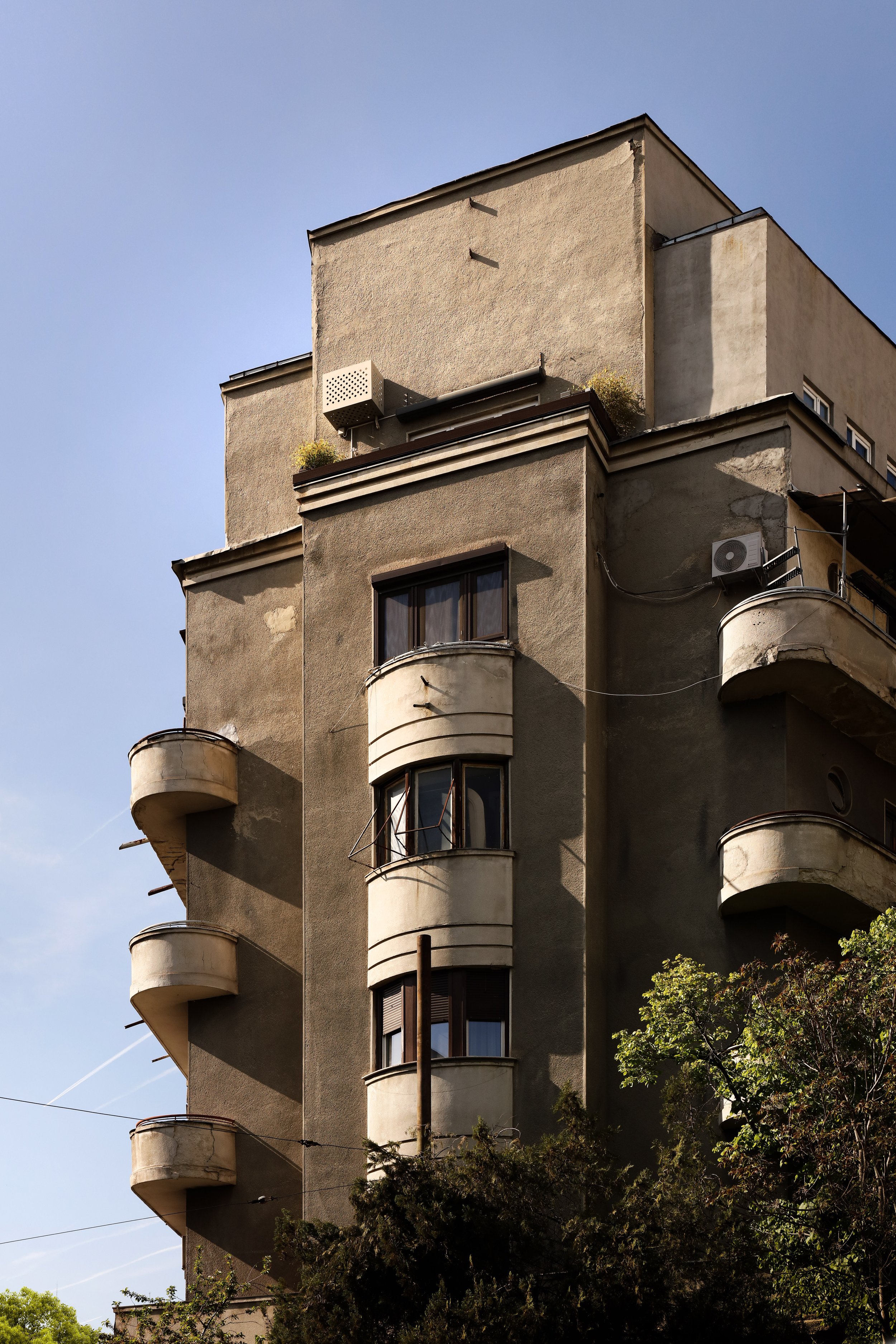 A tall apartment building with curved balconies and beige exterior walls, with some windows visible, set against a clear blue sky. There is a tree in the foreground.
