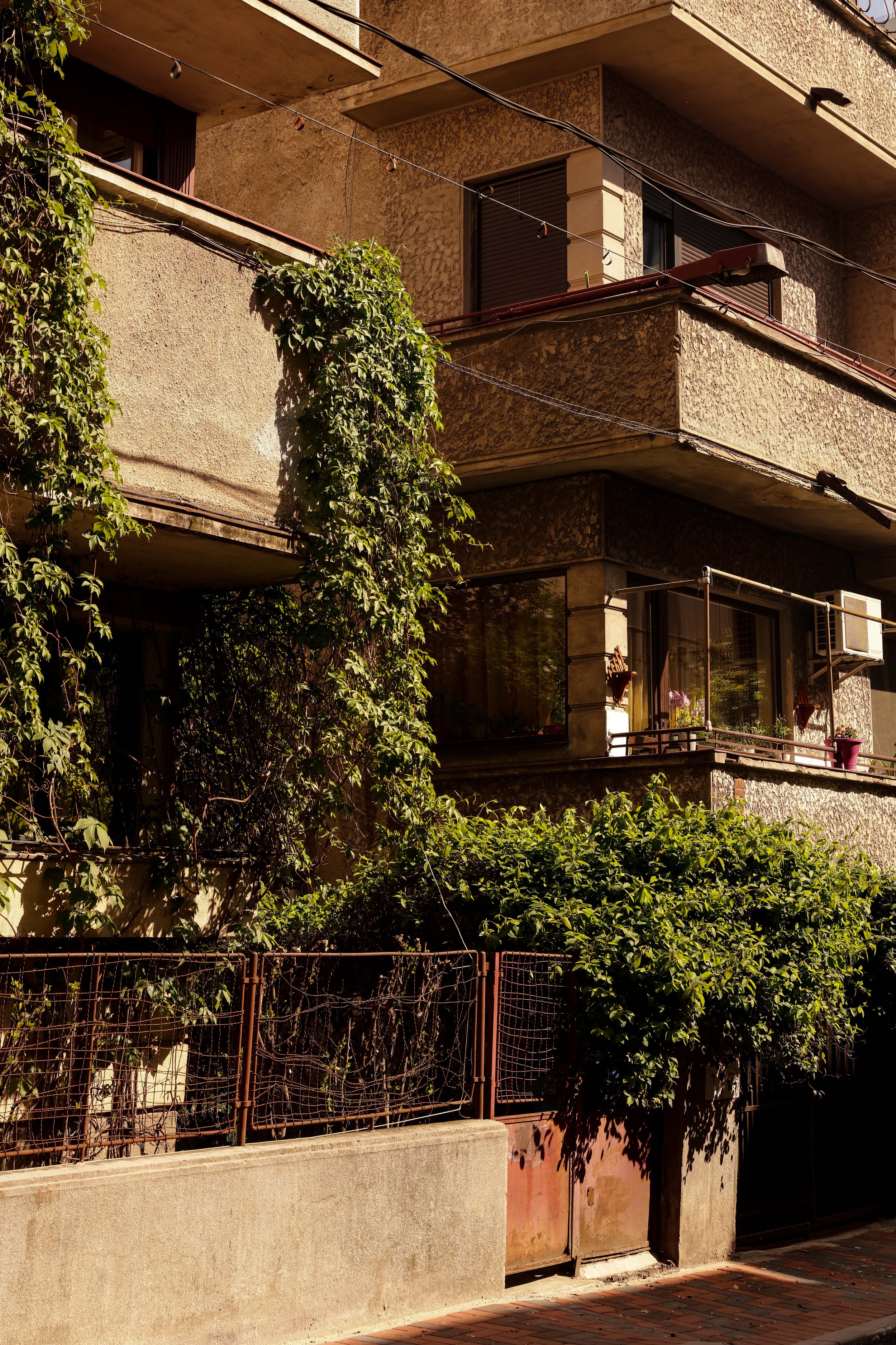 Old apartment building with balconies, closed shutters, some plants, and a rusty gate. The facade is textured concrete in beige and brown tones.