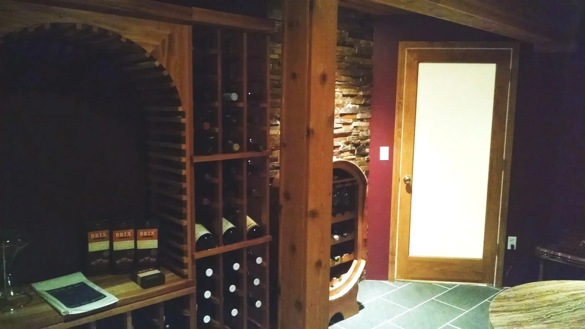 A wine cellar with wooden wine racks, bottles, and a stone accent wall, next to a door with a frosted glass panel.