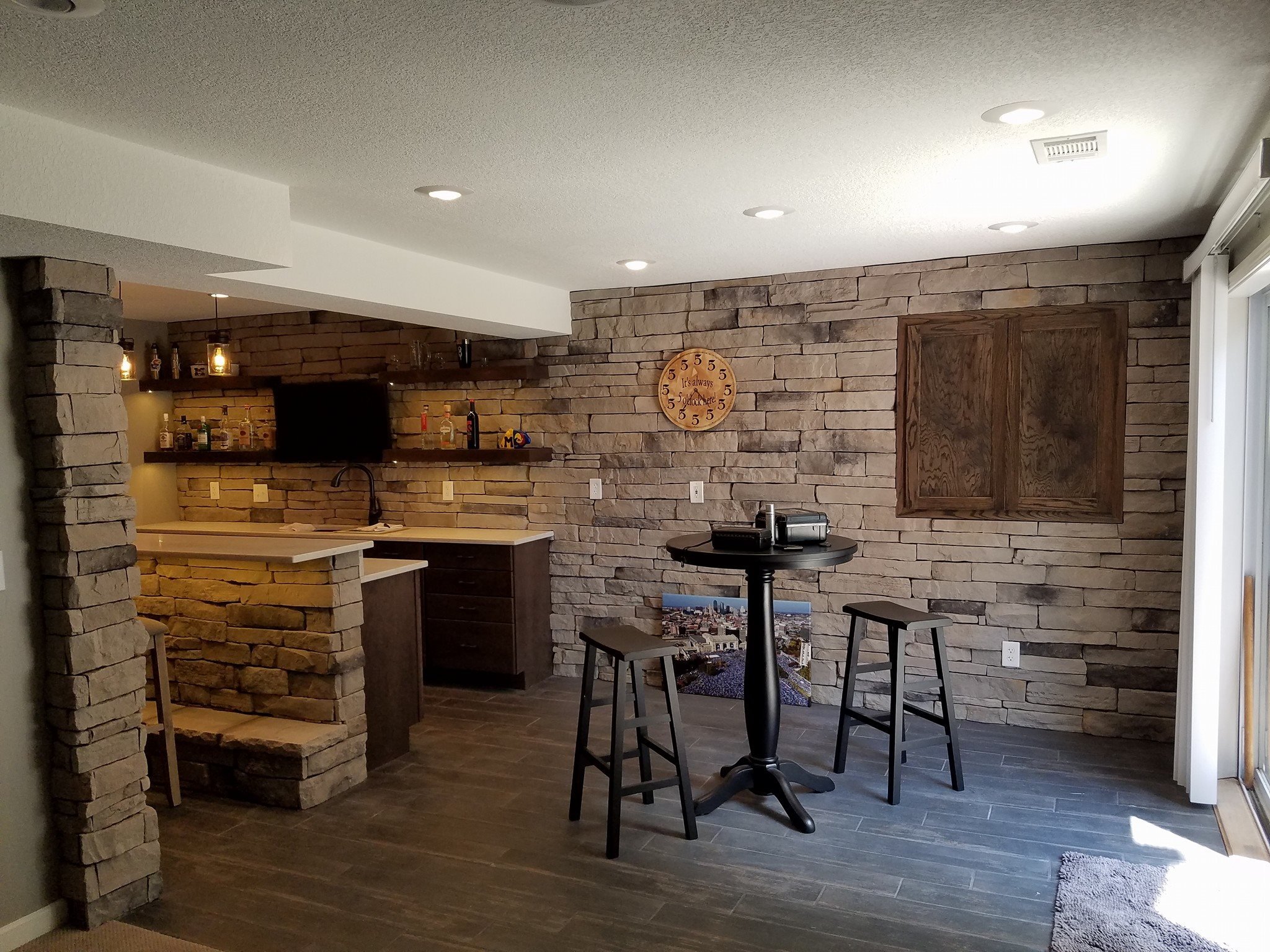 A cozy basement bar with a stone accent wall, a small tasting area with round table and two chairs.  A mounted flat screen TV, dart board and a bar area for entertaining.