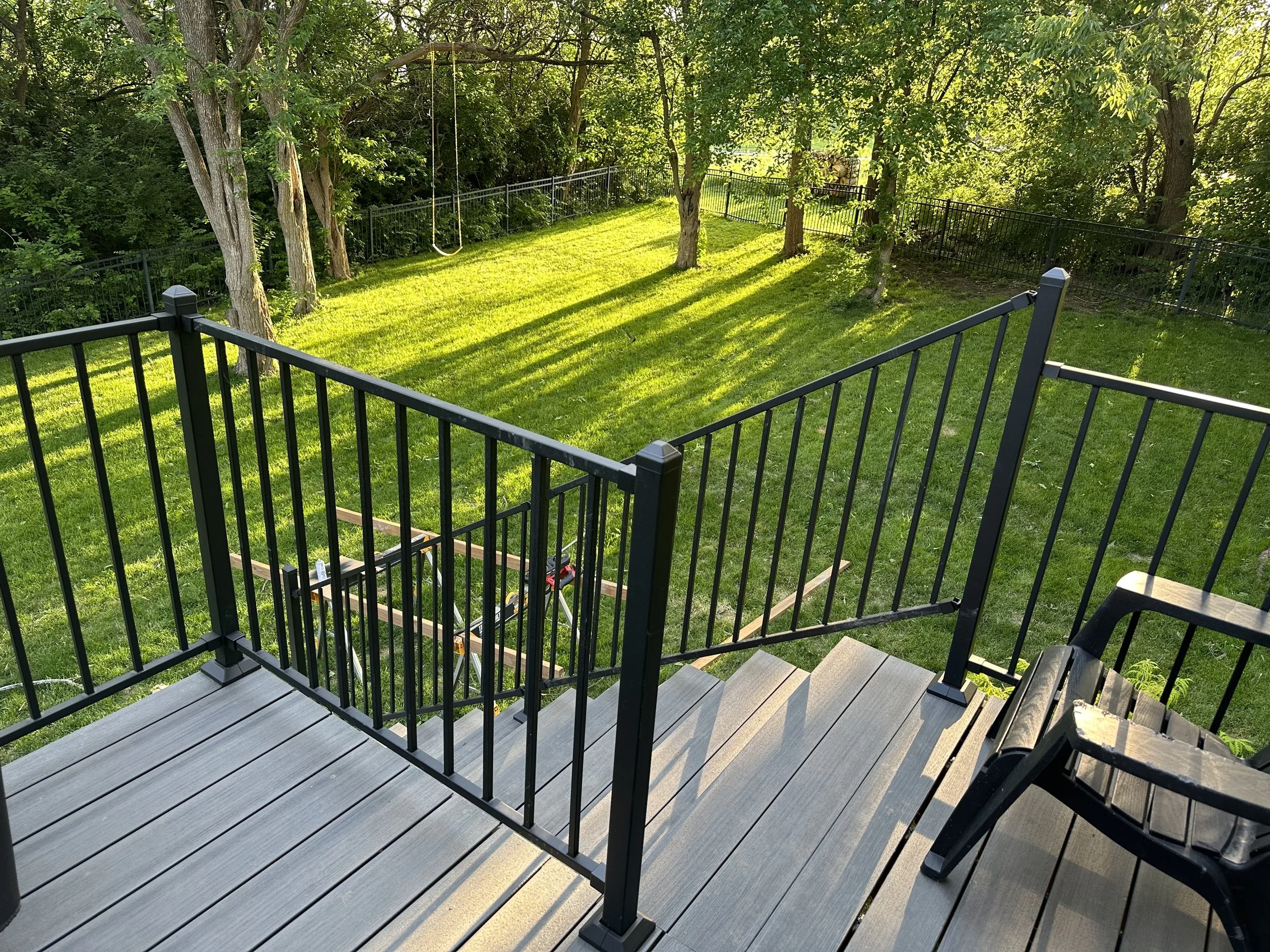 View from a newly completed composite deck with black metal railing, overlooking a grassy backyard with trees, a swing, and a fenced area in the sun.