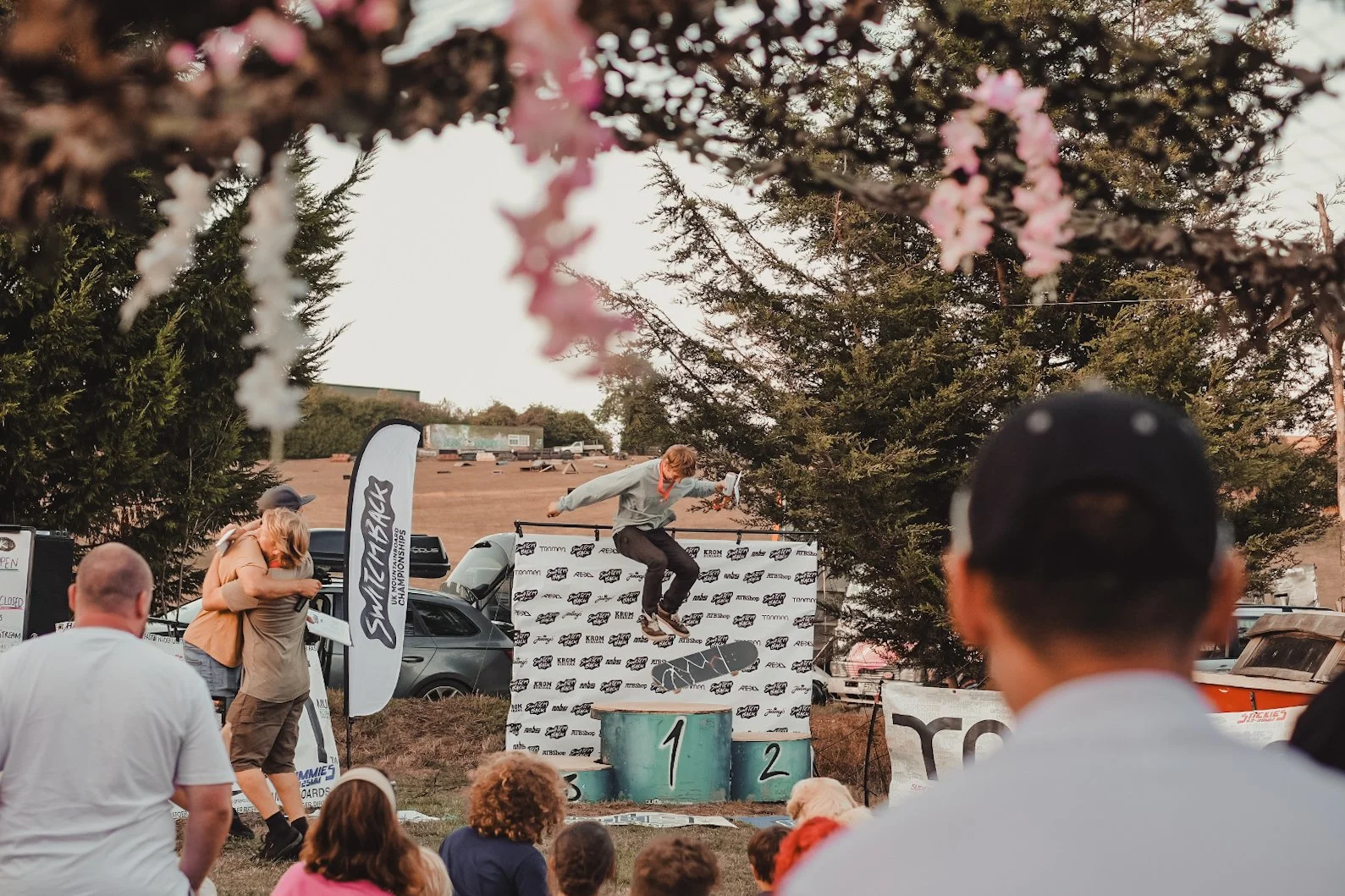 A skateboarder performing a trick on a podium at an outdoor skateboarding competition, with spectators watching and hugging, dressed casually, and trees and cars in the background.
