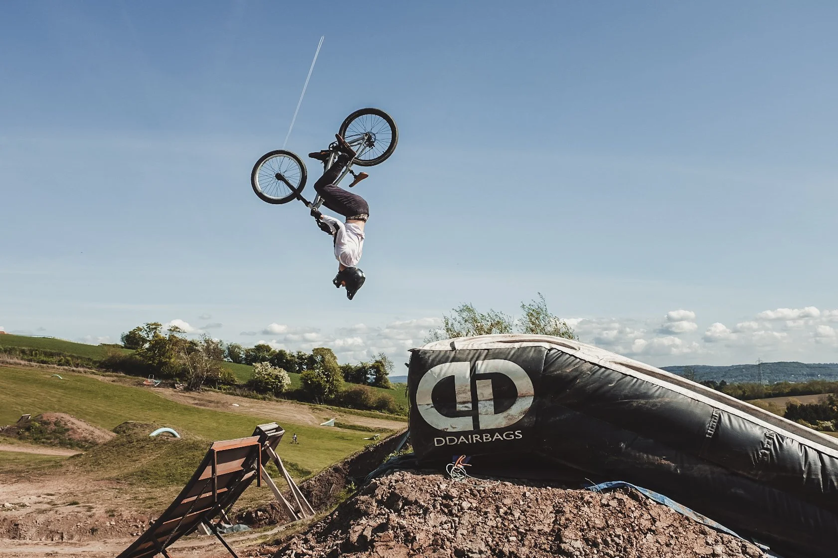 A person performing a flip on a BMX bike in mid-air at an outdoor dirt park, with clear blue sky and green hills in the background.
