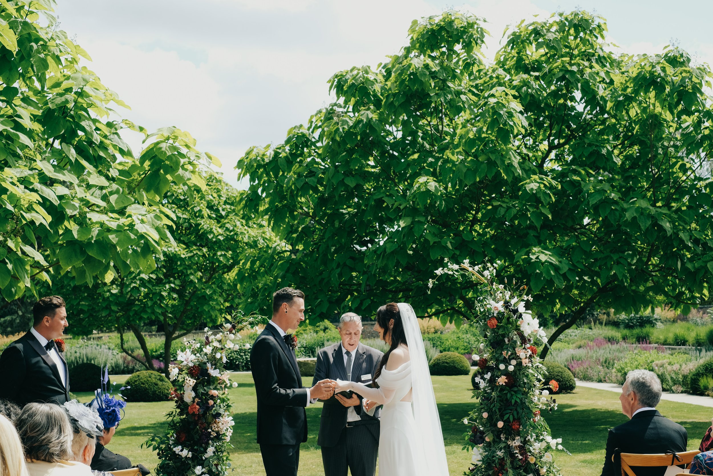 A wedding ceremony outdoors in a garden with tall green trees, where a bride and groom are exchanging rings under an arch decorated with flowers, officiated by an officiant. Guests are seated, witnessing the event.