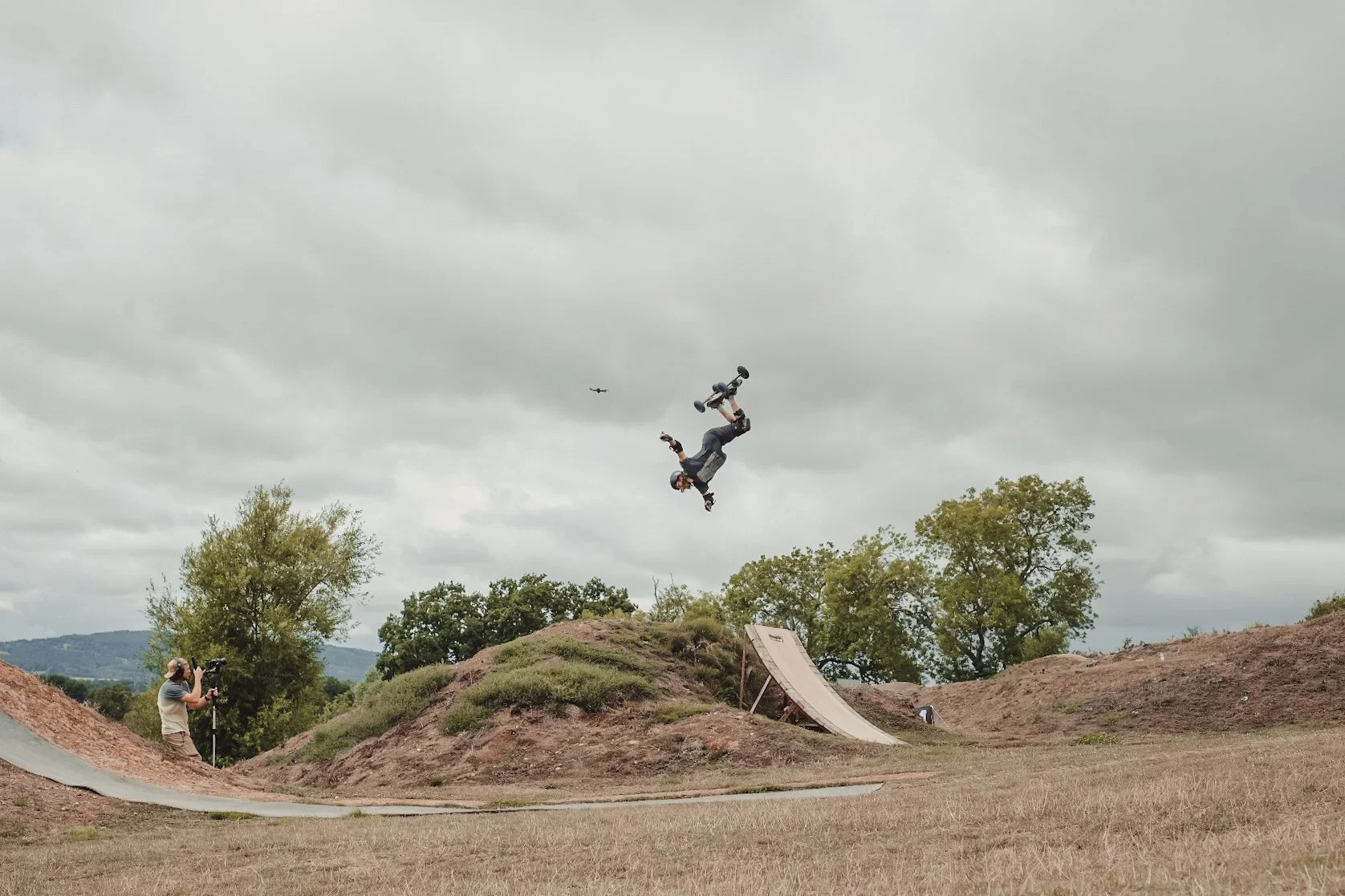 A person performing a high-flying stunt on a skateboard at an outdoor skate park with dirt mounds and a ramp, under a cloudy sky, while another person films the action.