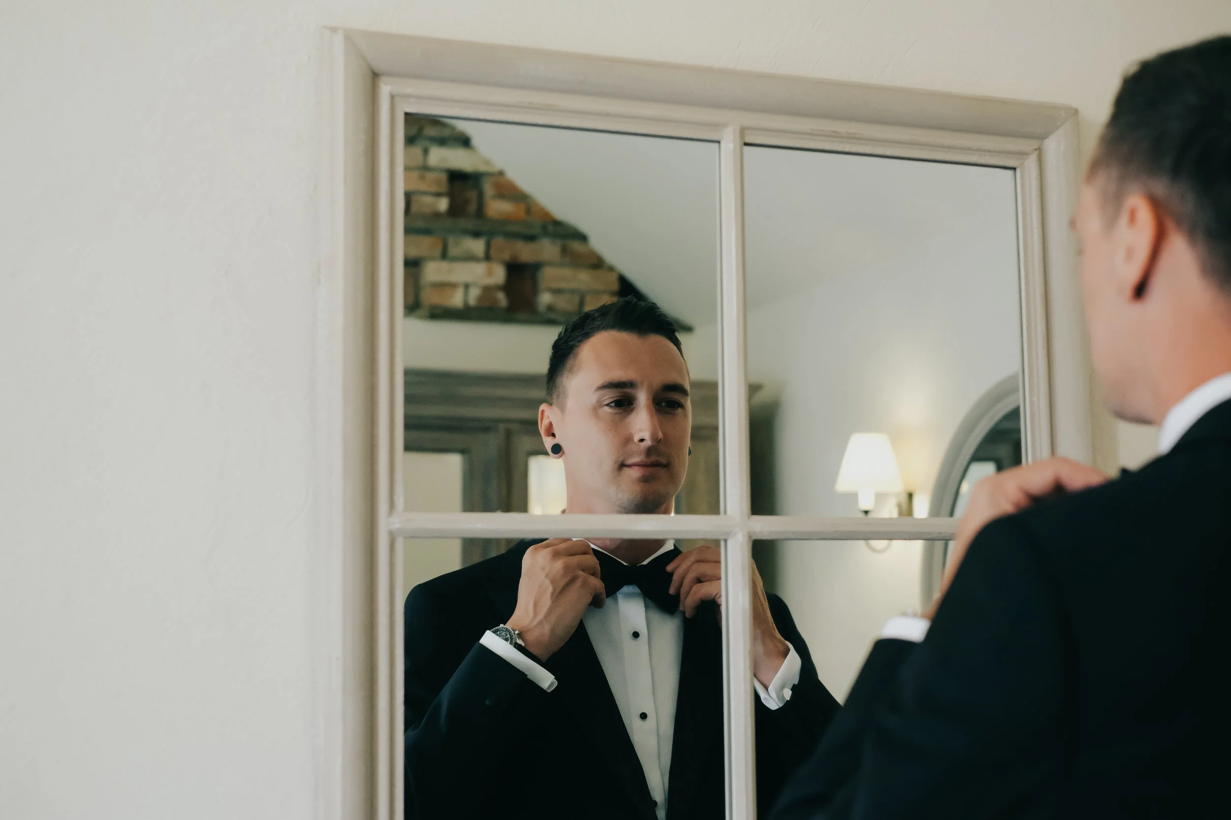 A man in a tuxedo adjusts his bow tie in front of a mirror, preparing for a formal event in a well-lit room.