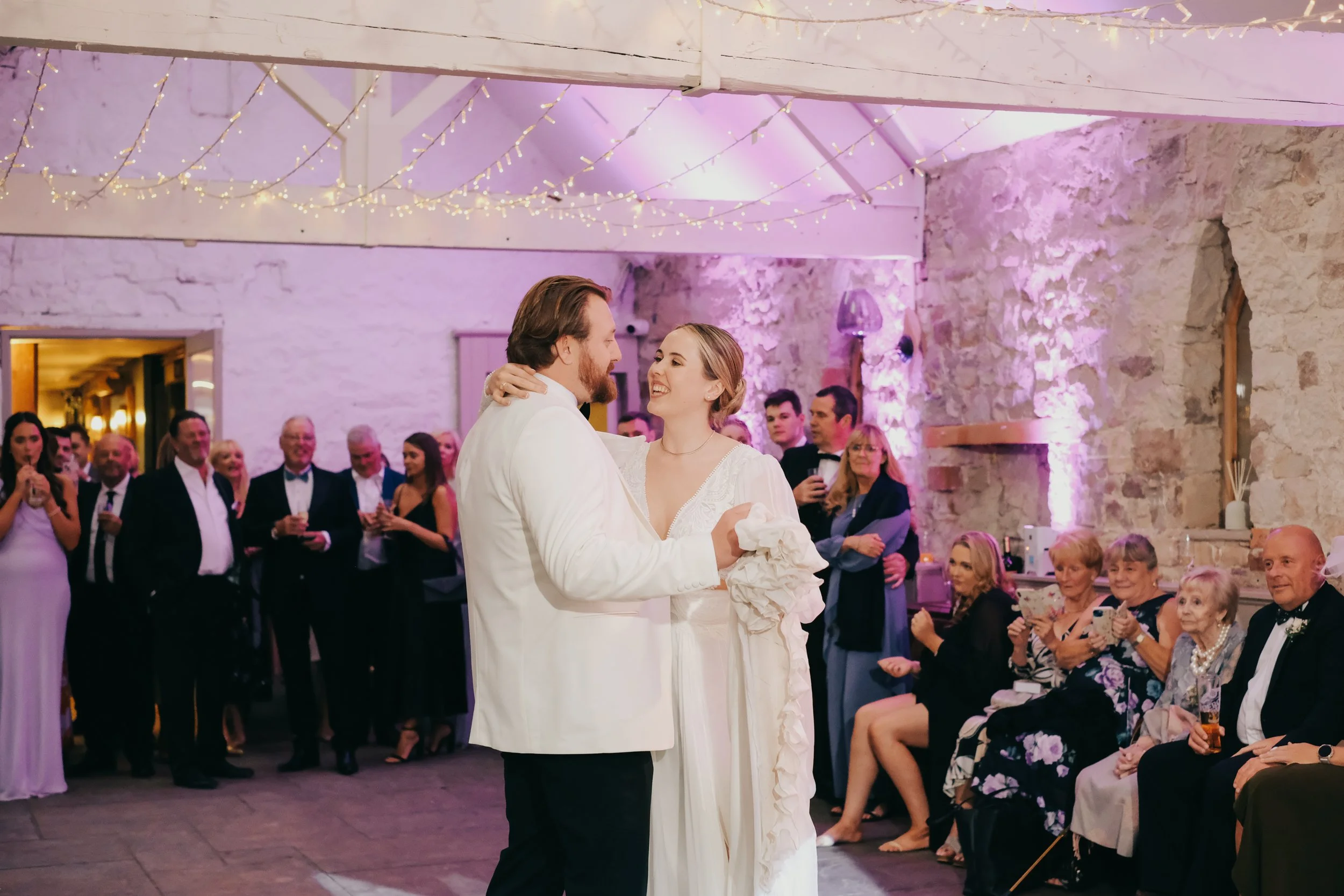 Bride and groom dancing at wedding reception in a rustic venue with stone walls and string lights, surrounded by seated and standing guests.