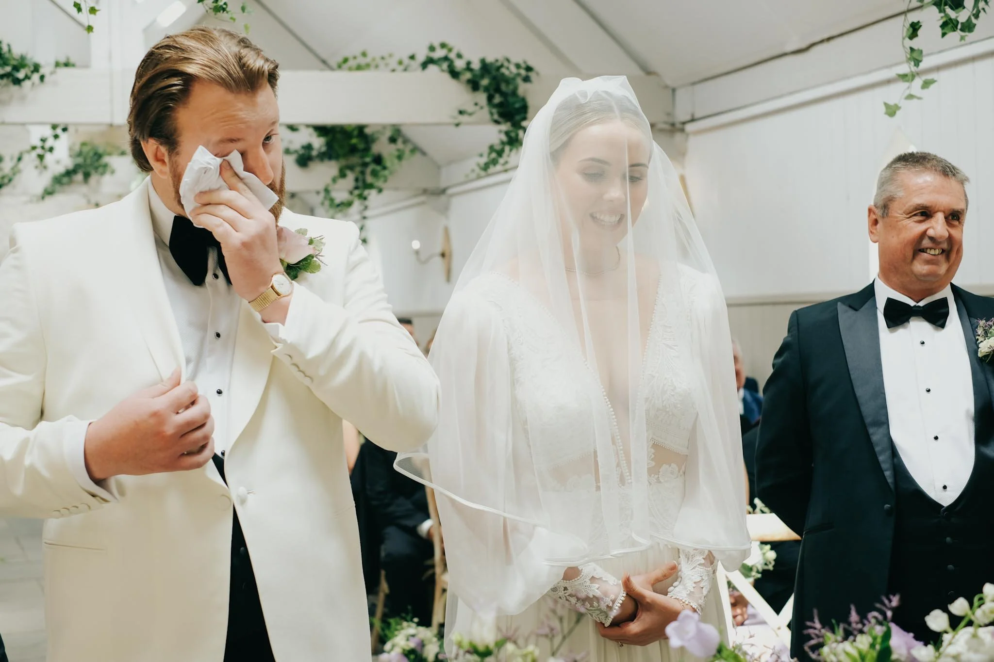 A bride and two men at a wedding ceremony, with the man on the left wiping tears with a tissue, the bride smiling under her veil, and the man on the right smiling, all dressed in formal wedding attire.
