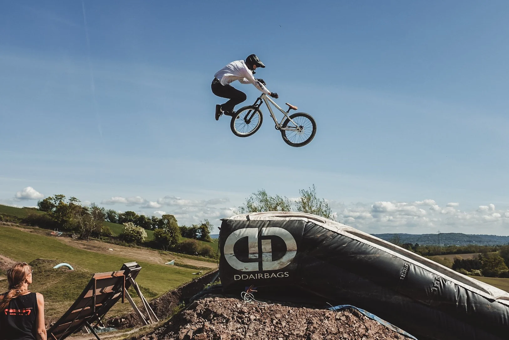 A person in a helmet and white shirt riding a mountain bike mid-air over a ramp at a dirt bike jump park with hills and trees in the background.