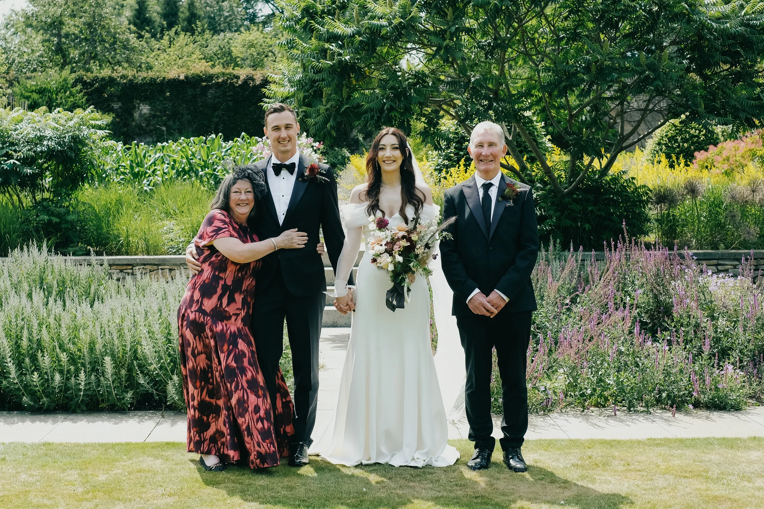 Group photo at a wedding in a garden with four people: a woman in a long red and black dress hugging a man in a tuxedo, a woman in a white wedding dress holding a bouquet of flowers, and a man in a black suit, all smiling.