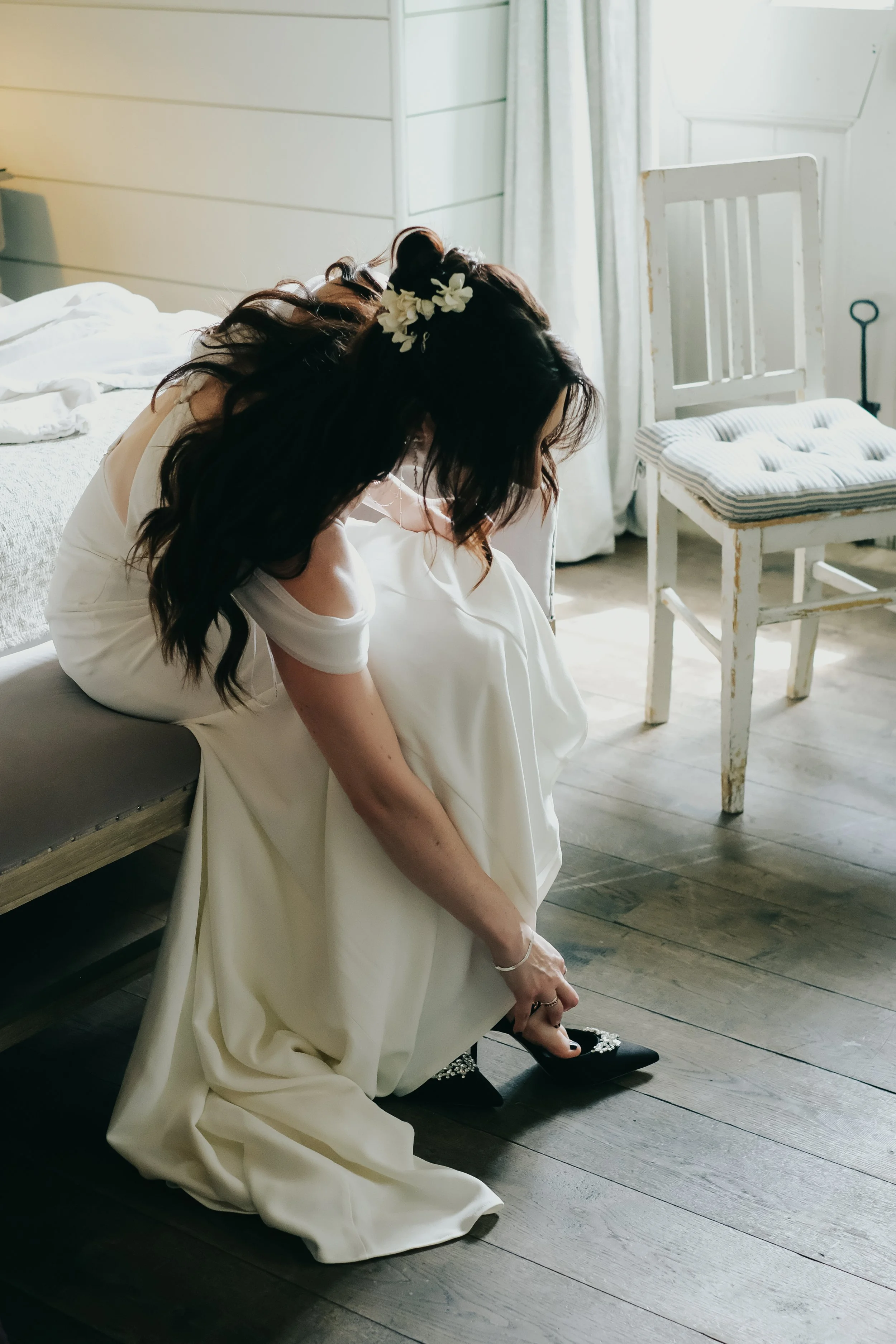 A woman in a white dress sits on a bench, tying her black high-heeled shoe, with a floral hair accessory and dark wavy hair, in a room with white wooden walls, a bed, and a white wooden chair.