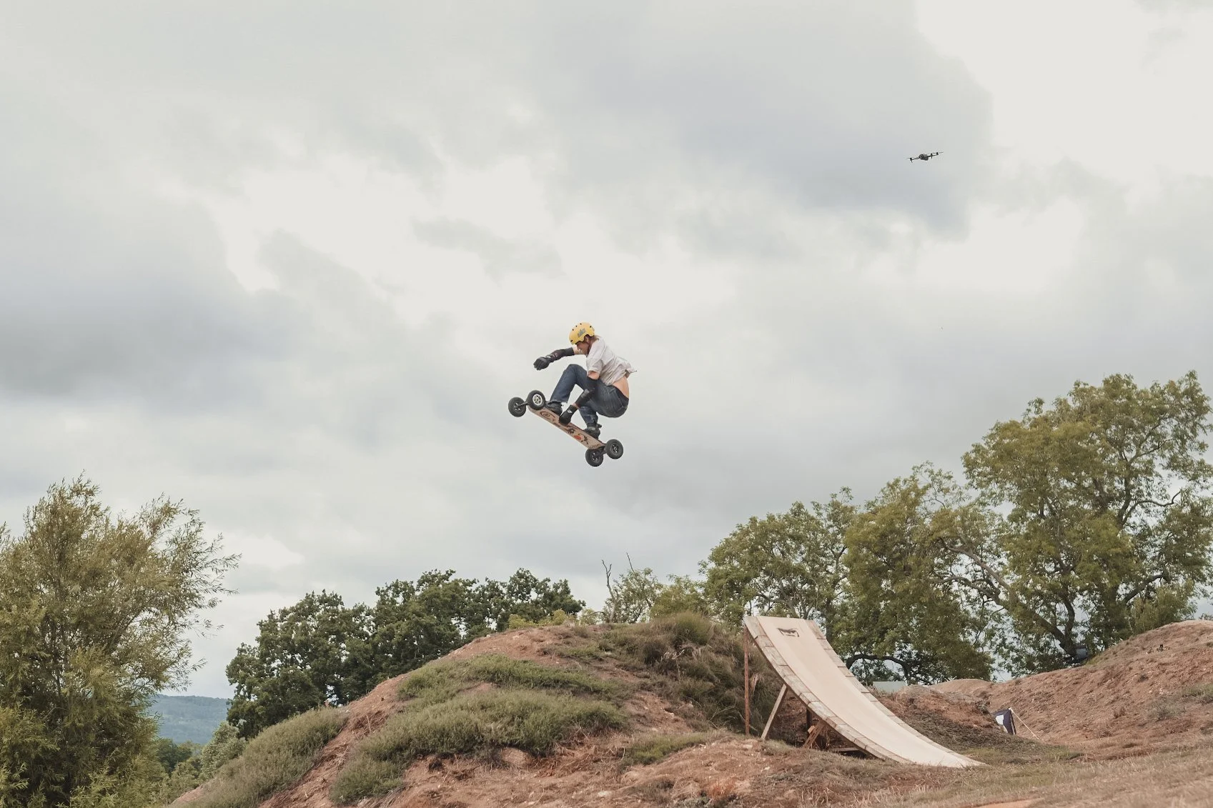 A person wearing a helmet is mid-air on a skateboard jumping off a ramp in an outdoor skate park with trees and cloudy sky in the background.