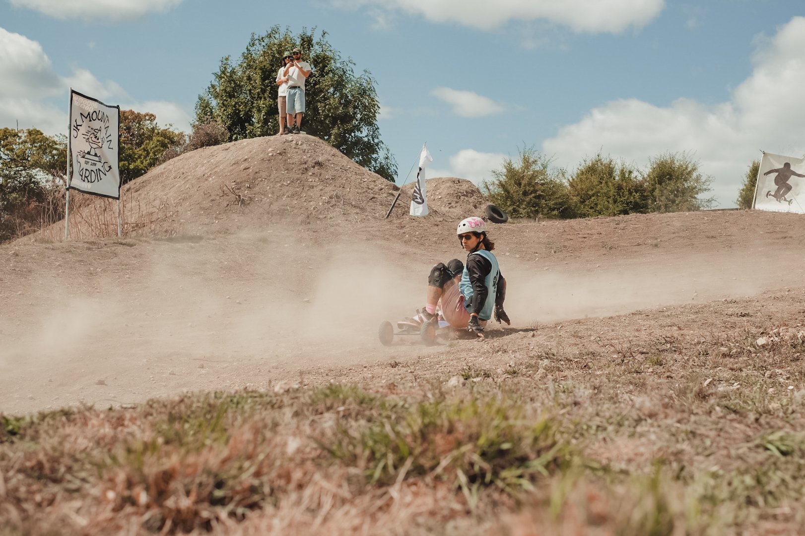 A person in protective gear and helmet has fallen off a skateboard on a dirt hill during a skateboarding event, with two spectators standing on top of the hill taking photos, and flags and signs in the background.