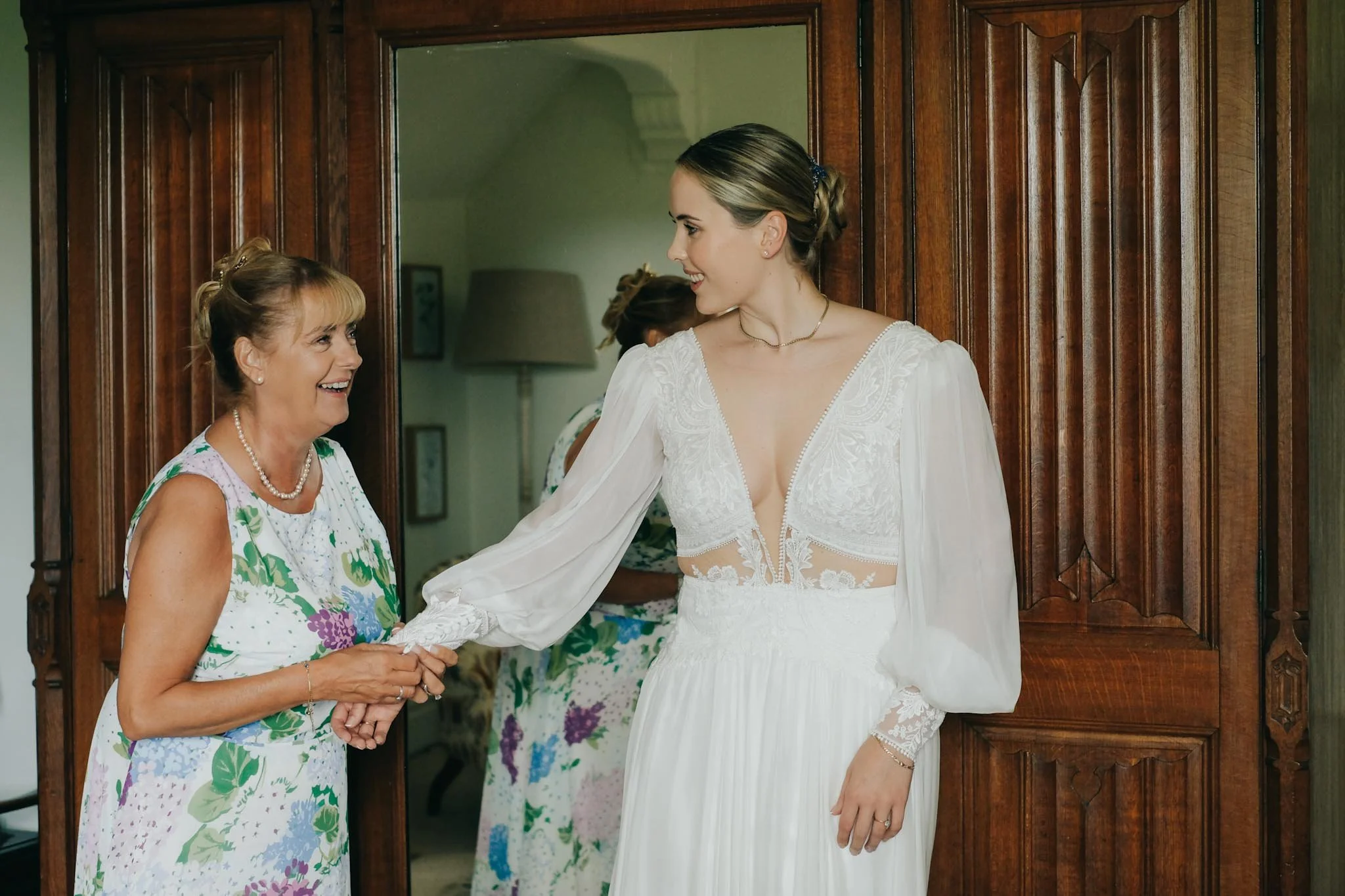 A bride wearing a white wedding dress with lace details and long puff sleeves smiling at an older woman in a floral dress, holding hands in a room with wooden furniture.