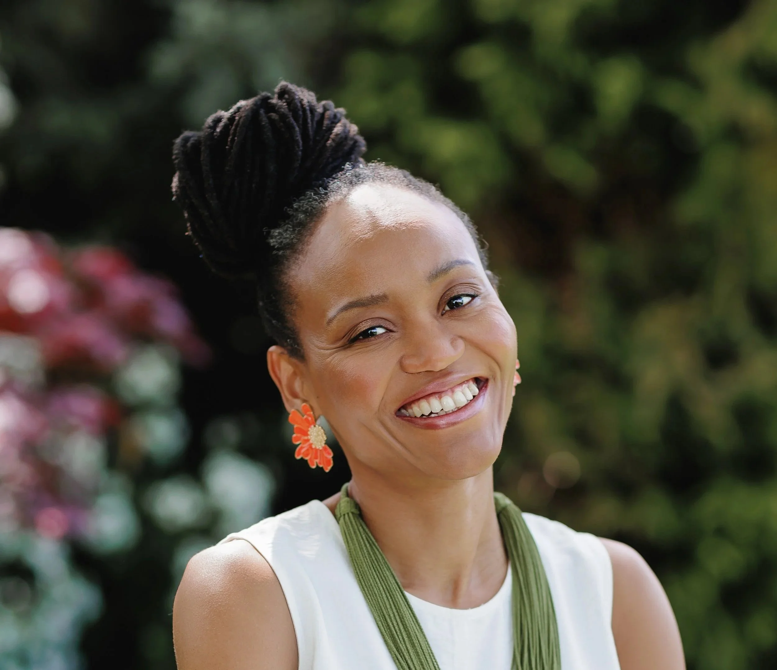 Smilin woman with updo hairstyle, orange flower earrings, and green necklace outdoors, background of greenery and burgundy flowers.