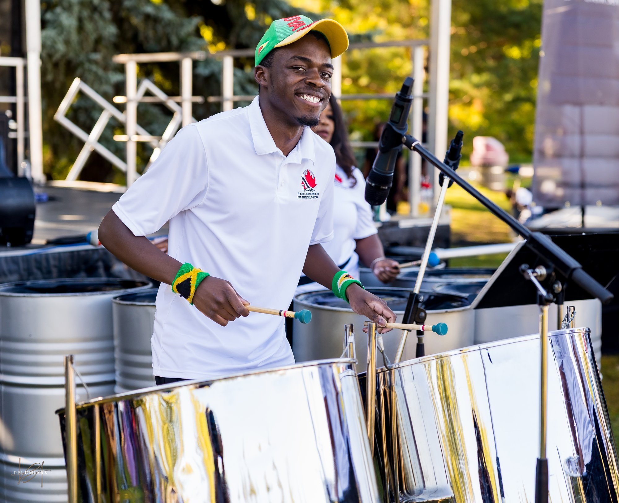 A young man playing steel drums outdoors during daytime with a big smile, wearing a white shirt with a logo, colorful wristbands, and a cap, with a stage and other musicians in the background.