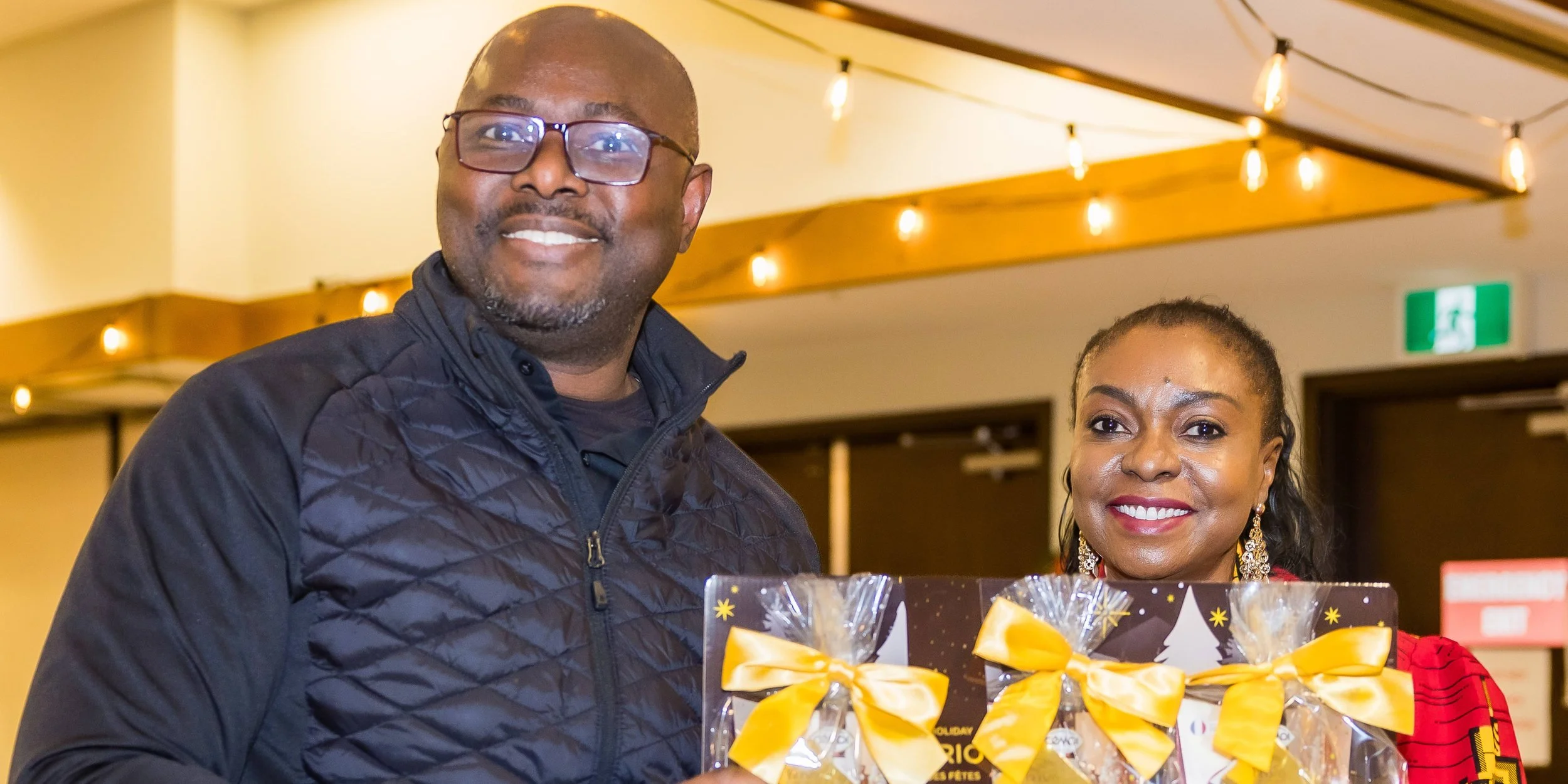A smiling man wearing glasses and a black jacket standing next to a smiling woman with earrings, holding a gift basket decorated with yellow ribbons and holiday-themed packaging in an indoor setting with string lights.