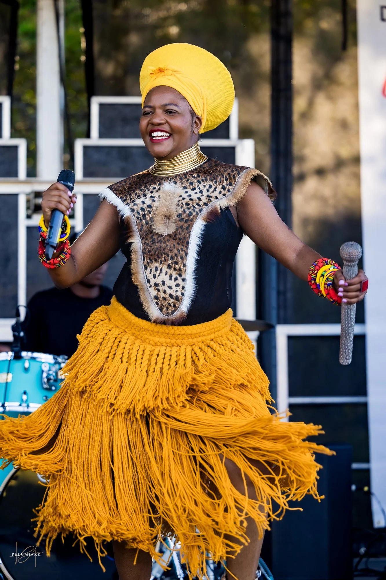 A woman dressed in vibrant African-inspired attire on stage, holding a microphone, smiling, wearing a yellow headwrap and skirt with fringe, and accessories, with musical equipment in the background.