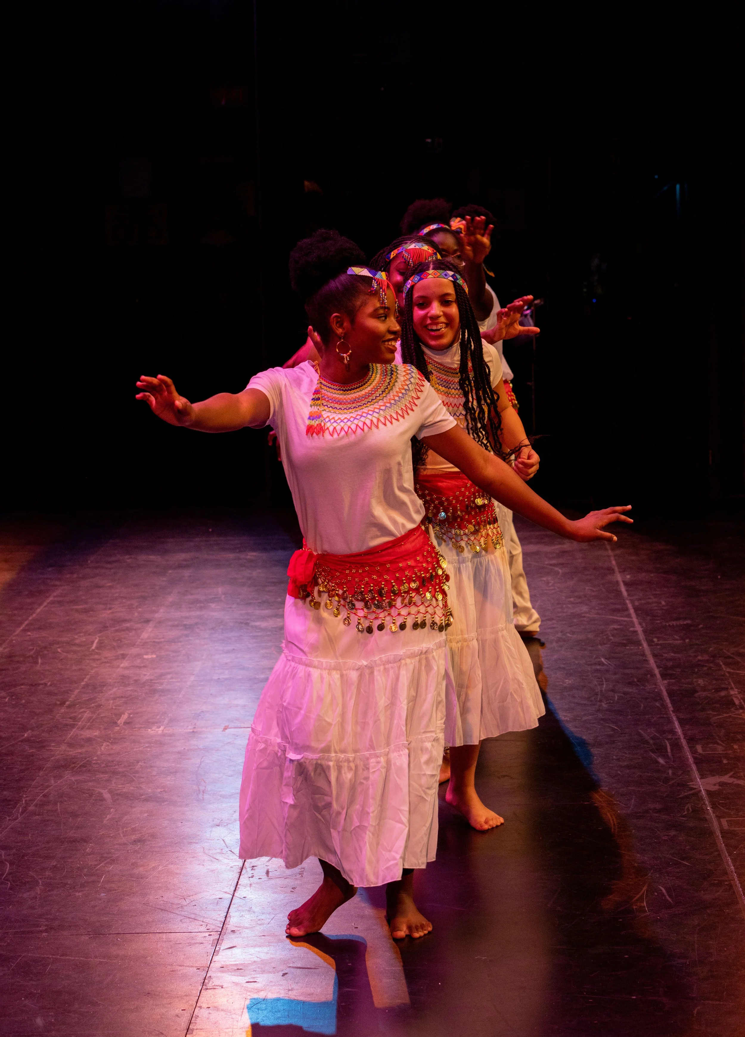 Group of women dancing on stage wearing traditional costumes with colorful beaded necklaces and headbands.