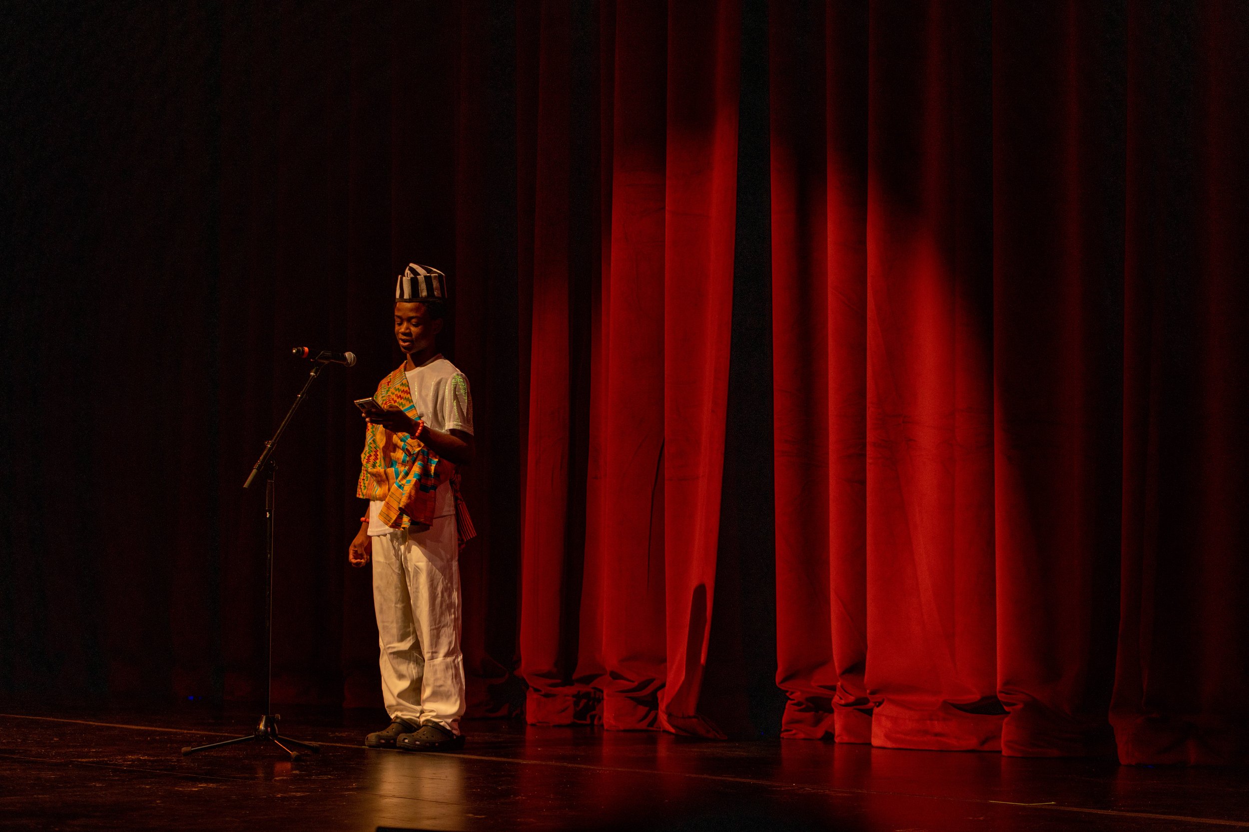 A person wearing traditional African attire stands on stage in front of red curtains, reading from a paper at a microphone.