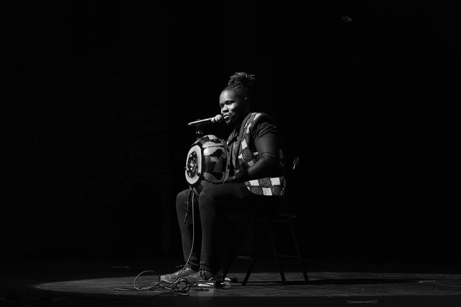 A person with dreadlocks sitting on a chair on a dark stage, playing a traditional African drum, with a microphone attached, in black and white.