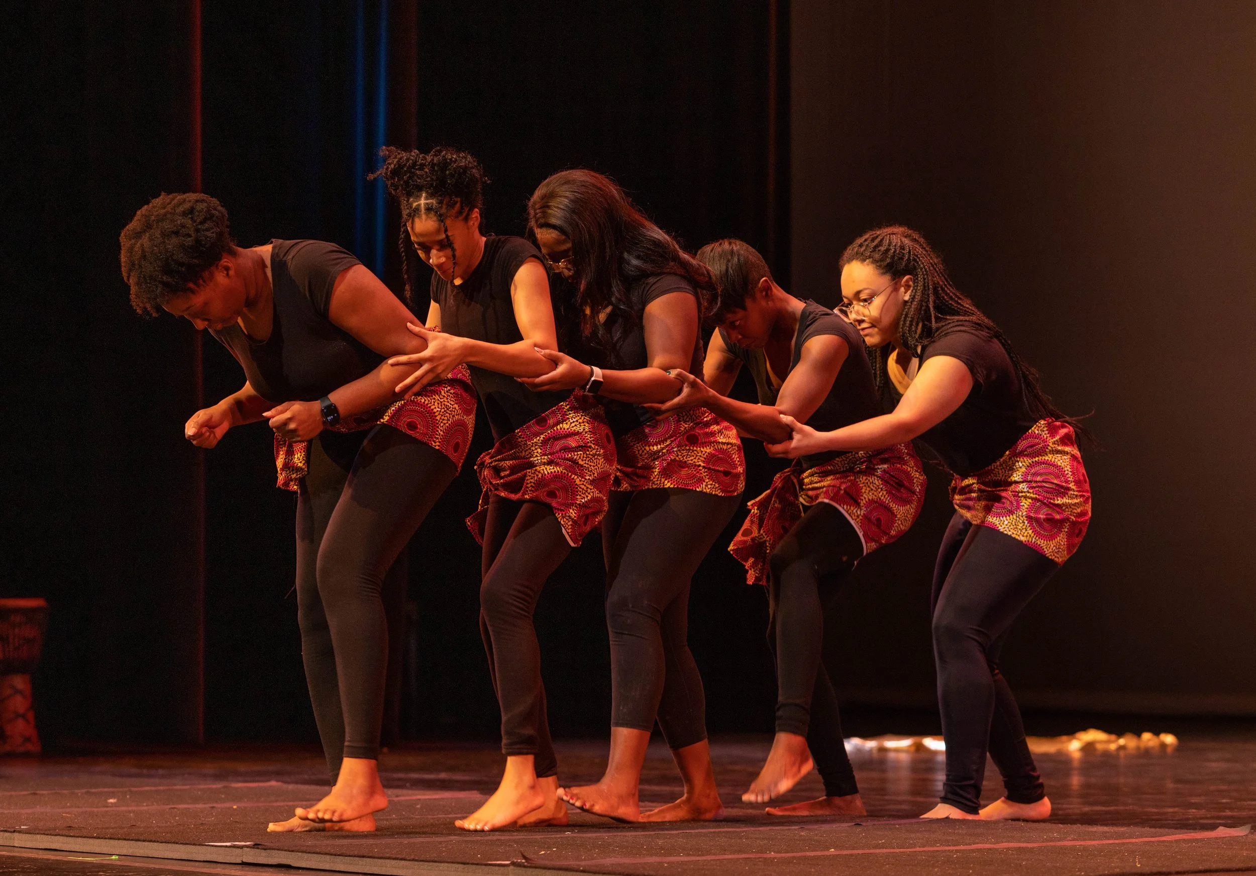 Group of five women performing a choreographed dance on stage, dressed in matching black shirts and red patterned skirts, holding each other's arms in a line.