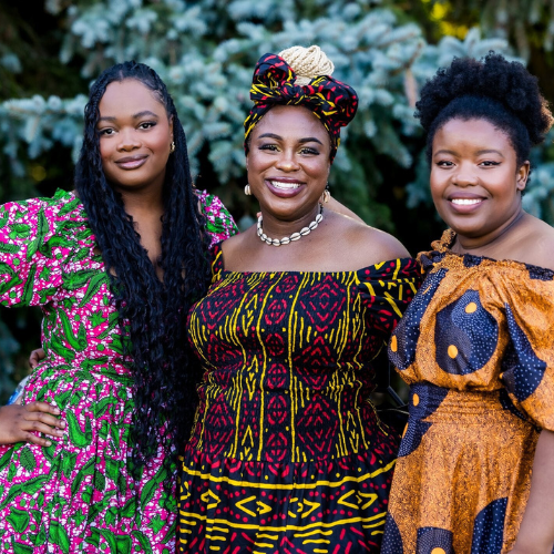 Three women dressed in colorful, traditional African prints standing outdoors in front of greenery, smiling at the camera.