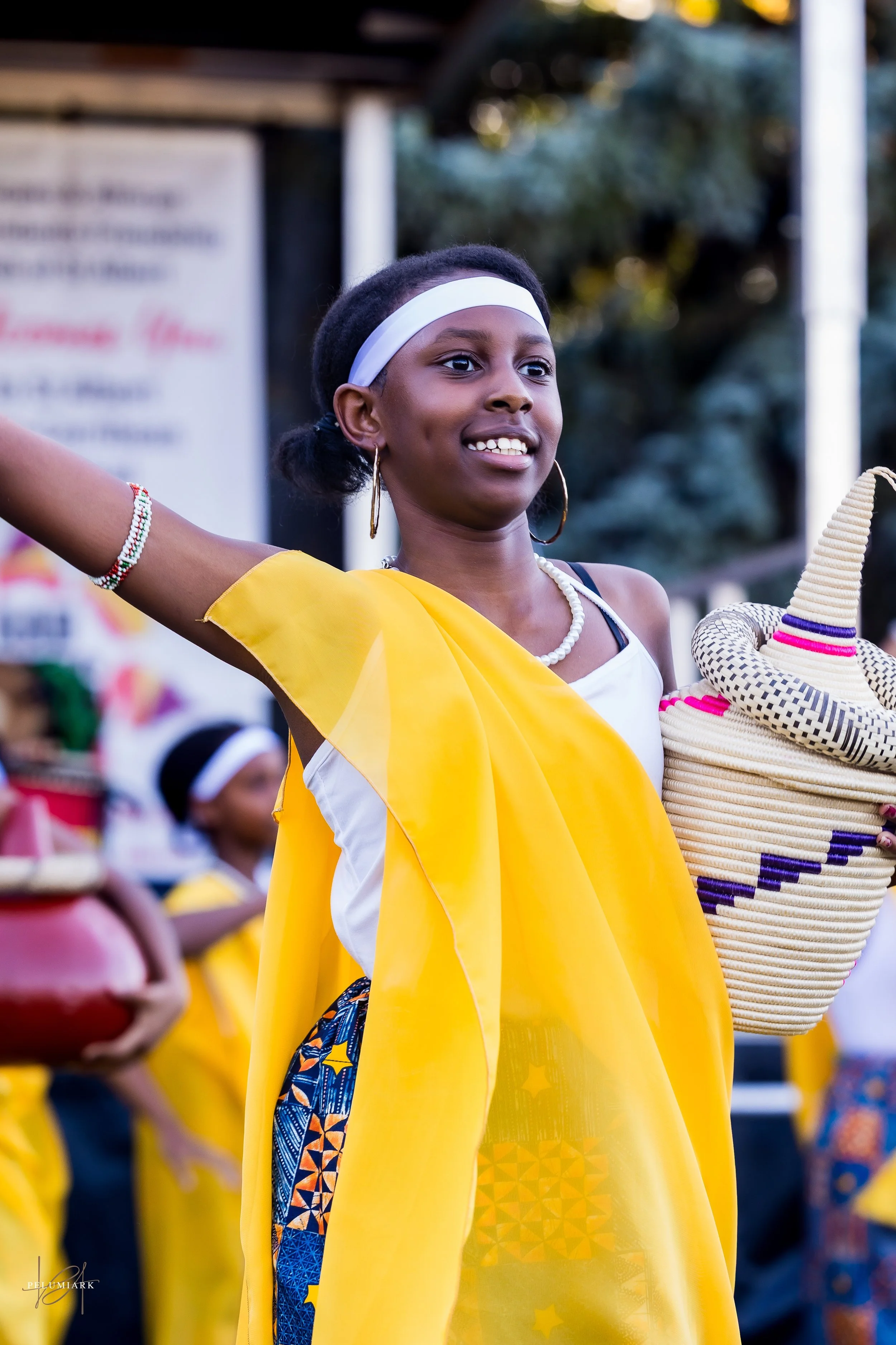 Young girl dressed in traditional colorful attire with a yellow shawl, holding a woven basket, smiling, and dancing outdoors during a cultural celebration.