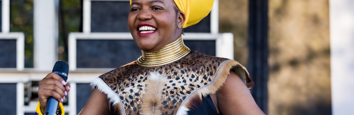 A woman wearing traditional African attire, including a yellow headwrap and a leopard print top, holding a microphone and smiling.