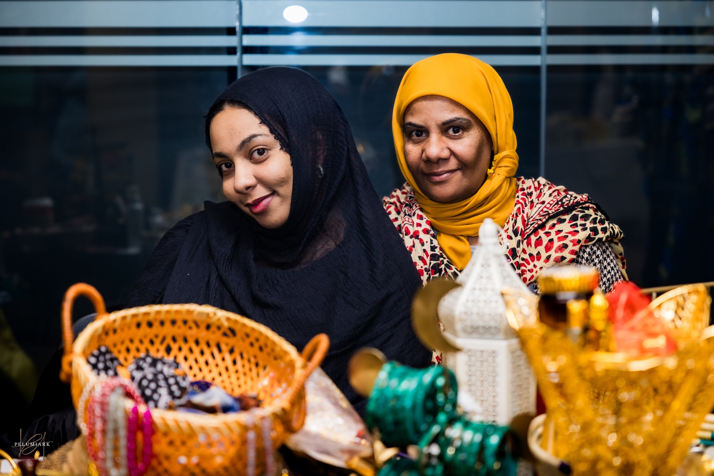 Two women wearing headscarves sitting behind a table with colorful jewelry and decorative items.