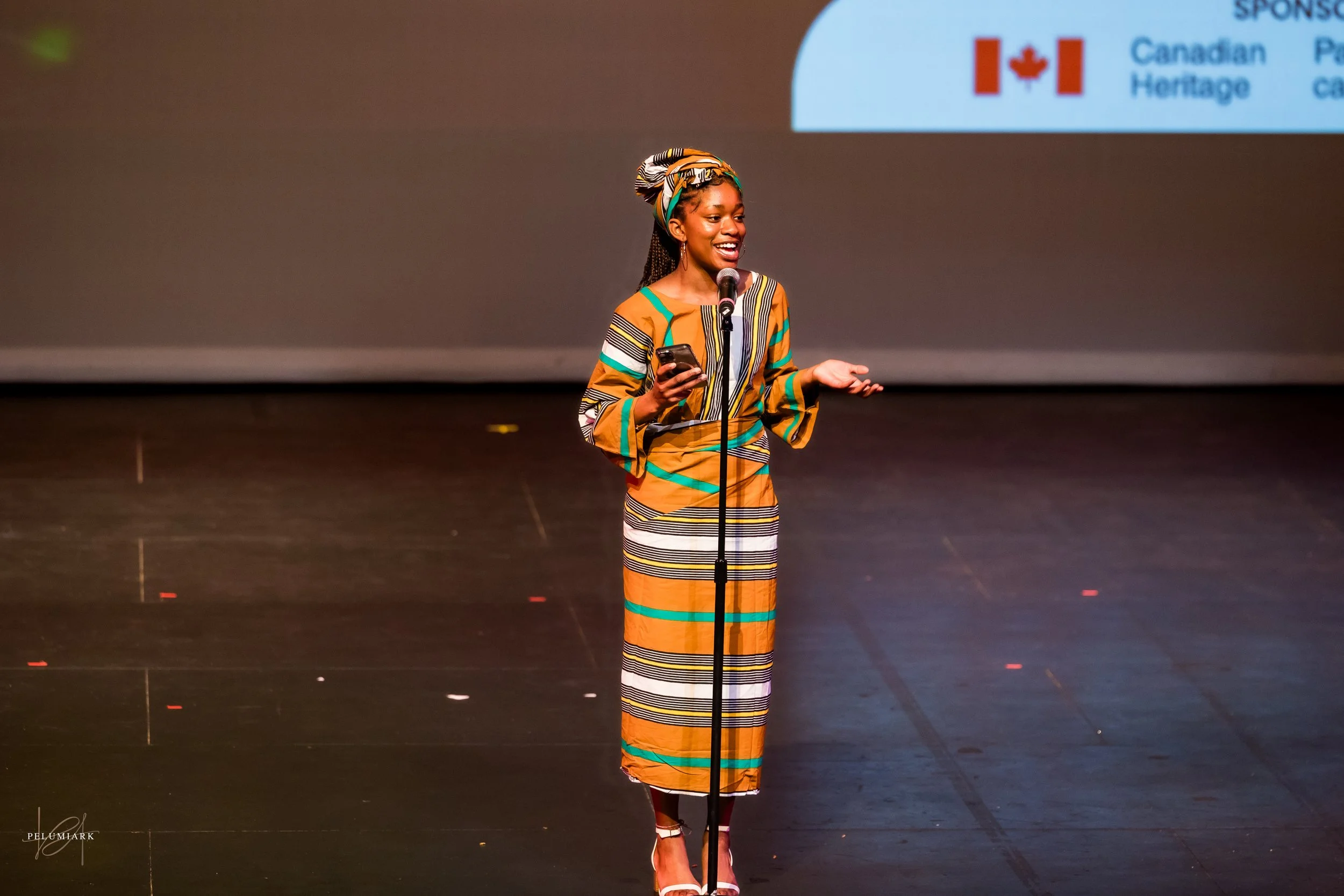 A woman wearing a colorful African print dress and headwrap is standing on a stage, speaking into a microphone, holding a phone in her left hand, and smiling.
