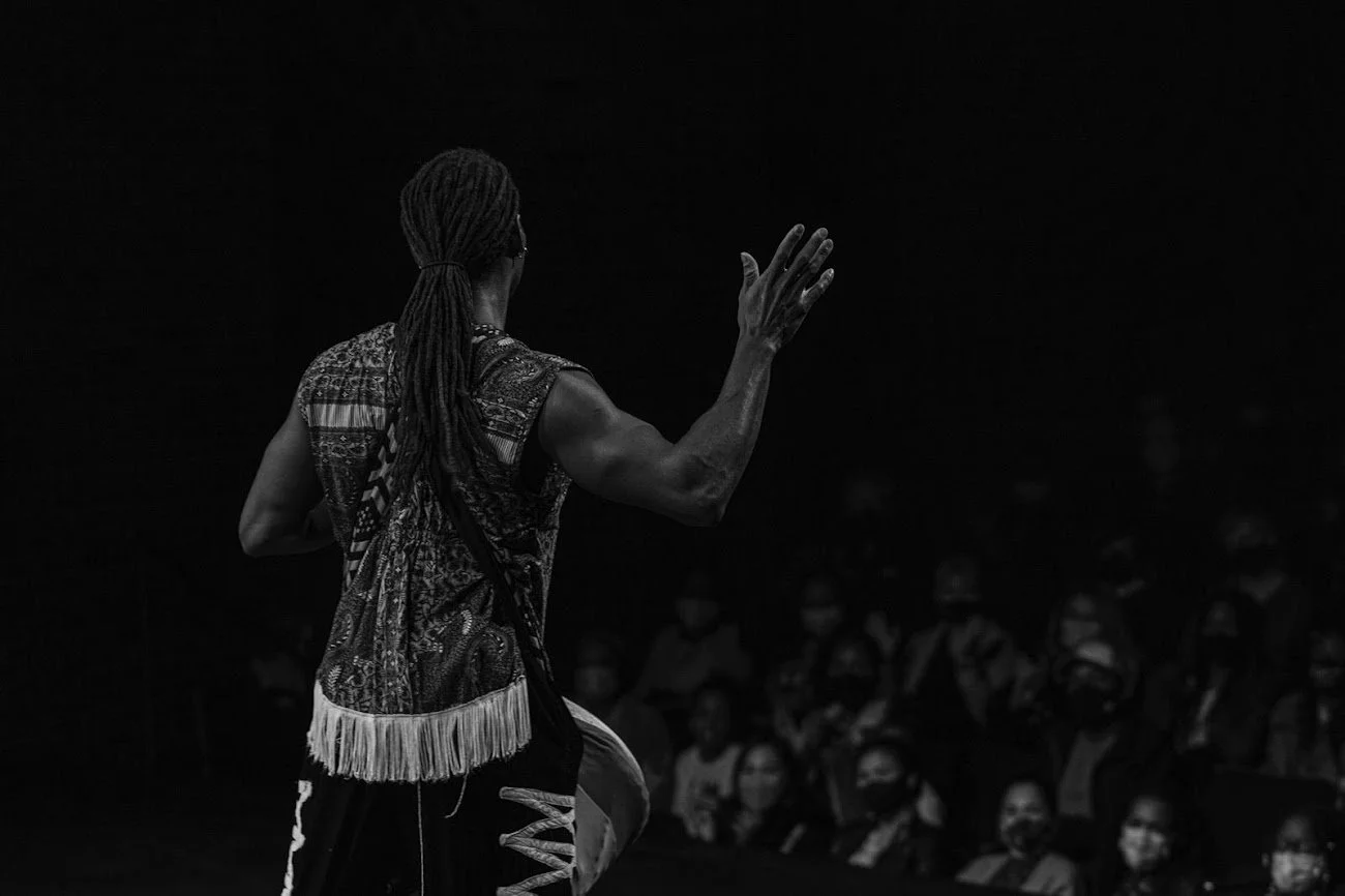 A woman with long braids gesturing with her hand on a dark stage, with an audience wearing masks in the background.