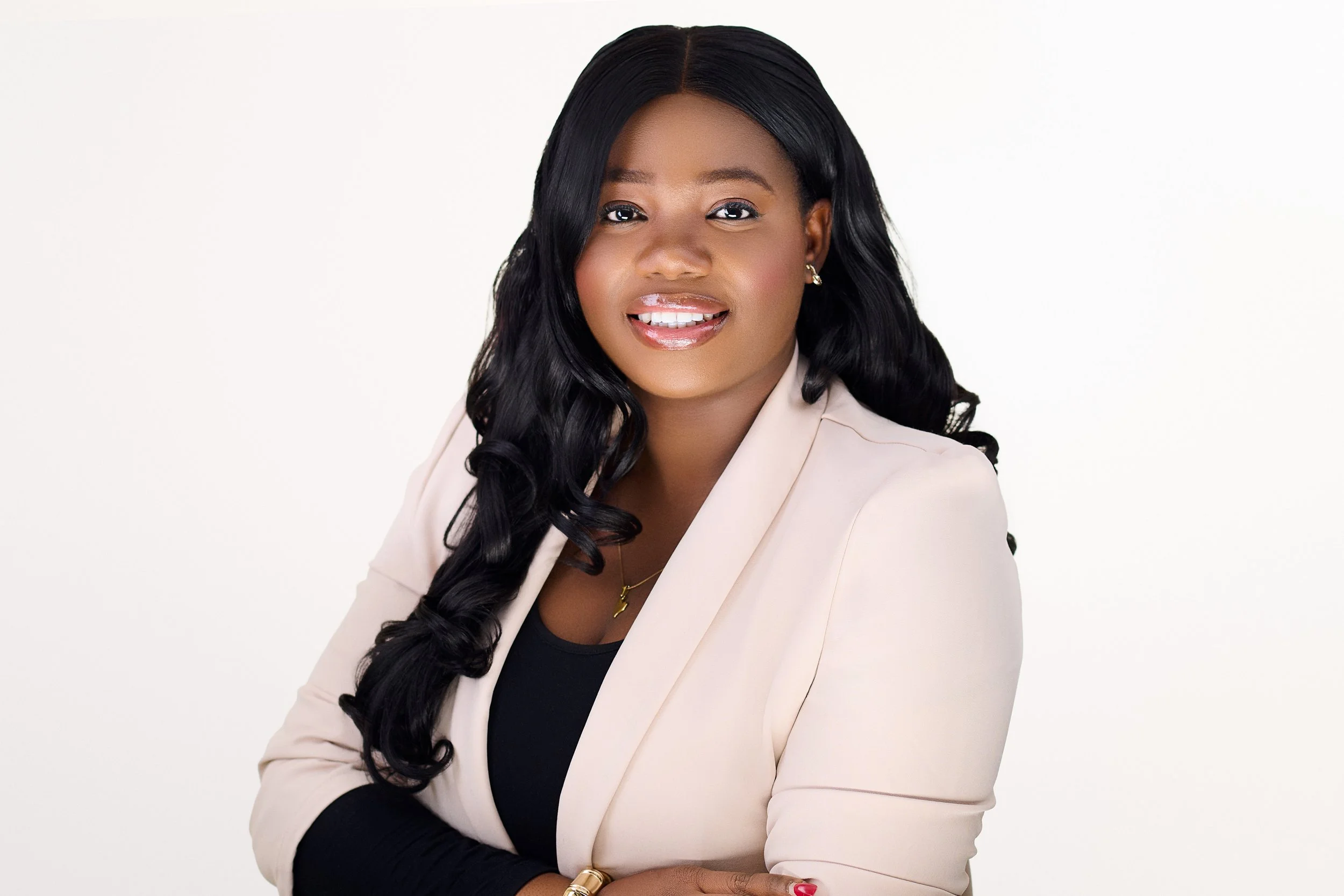 A smiling African American woman with long black wavy hair, wearing a light beige blazer over a black top, gold jewelry, and red nail polish, posed against a white background.