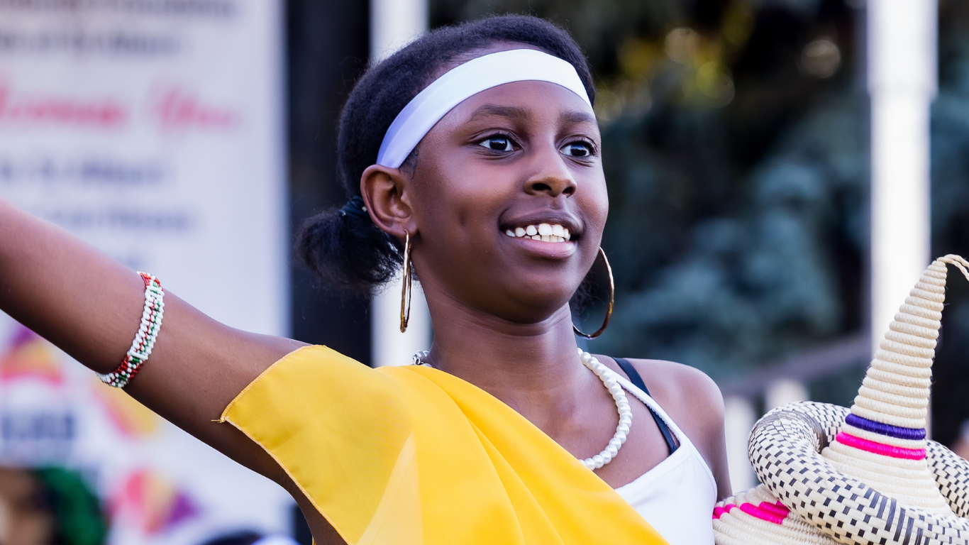 Young girl smiling, wearing a yellow shawl, white headband, hoop earrings, and beaded bracelet, holding a colorful woven hat with a conical shape, outdoors during the daytime.