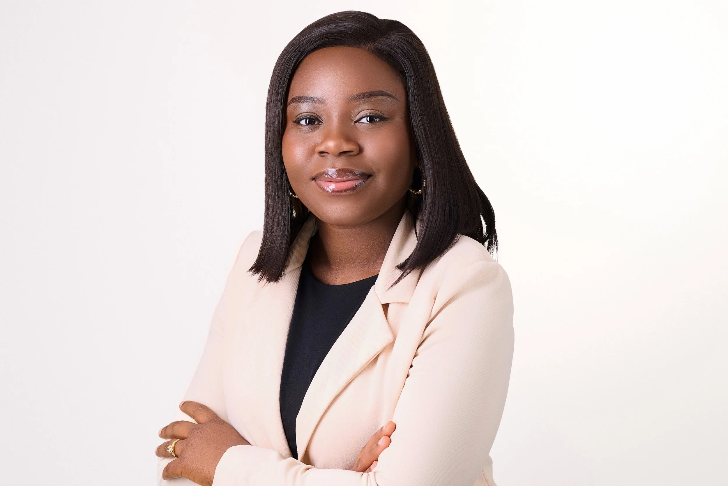 A young African American woman with shoulder-length straight black hair, wearing a beige blazer over a black top, crossing her arms, standing against a plain white background.