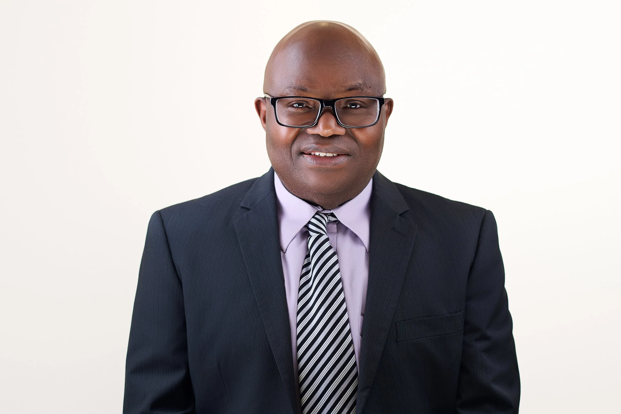 Professional portrait of a middle-aged African American man wearing glasses, a dark suit, a light purple shirt, and a striped tie, against a plain white background.