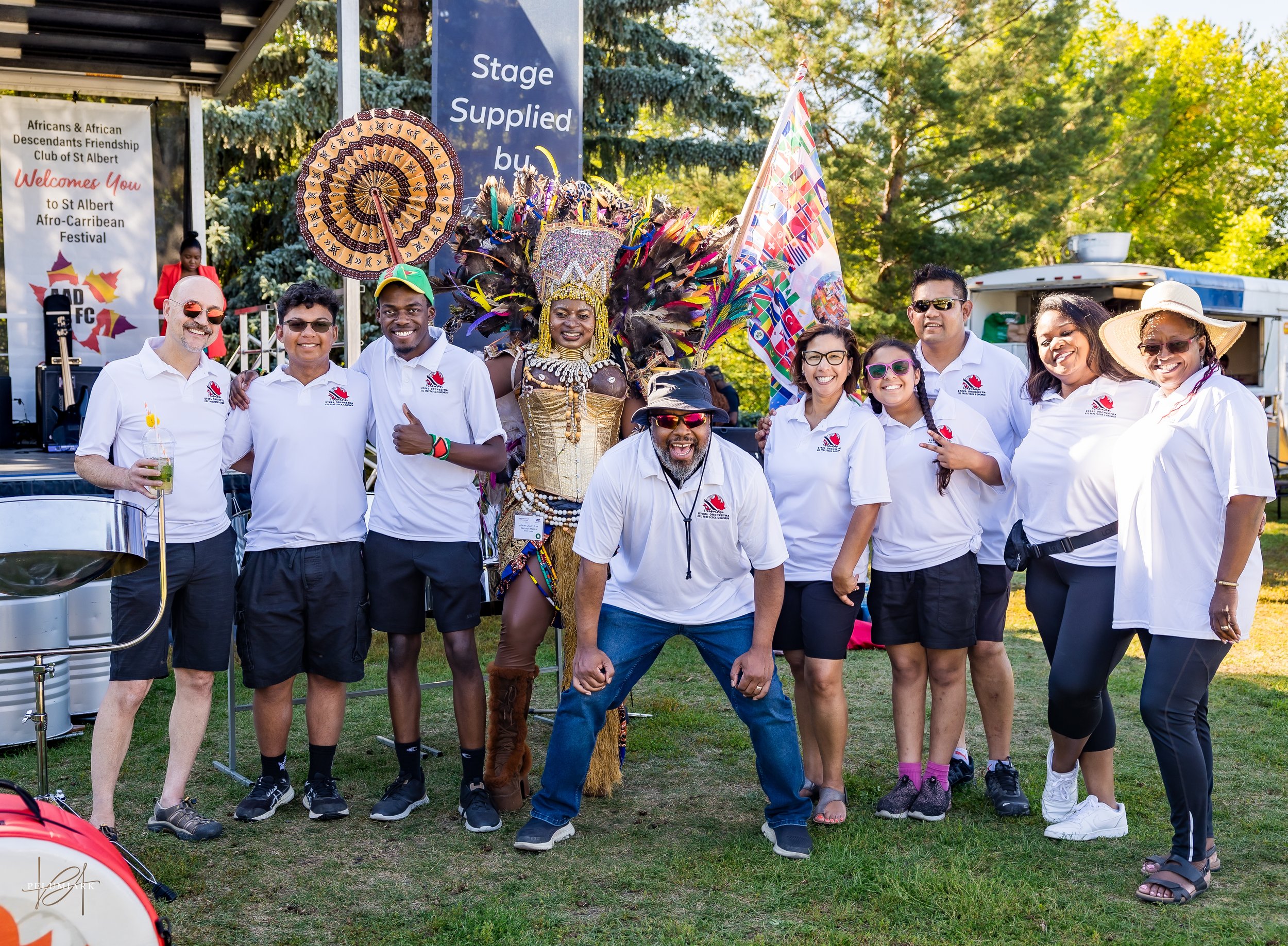Group of people standing outdoors in front of a festival stage, including a woman in traditional African attire with a large feathered headpiece, surrounded by men and women wearing matching white shirts from the African & African Descendants Friendship Club of St. Albert, celebrating at the St. Albert Afro-Caribbean Festival.