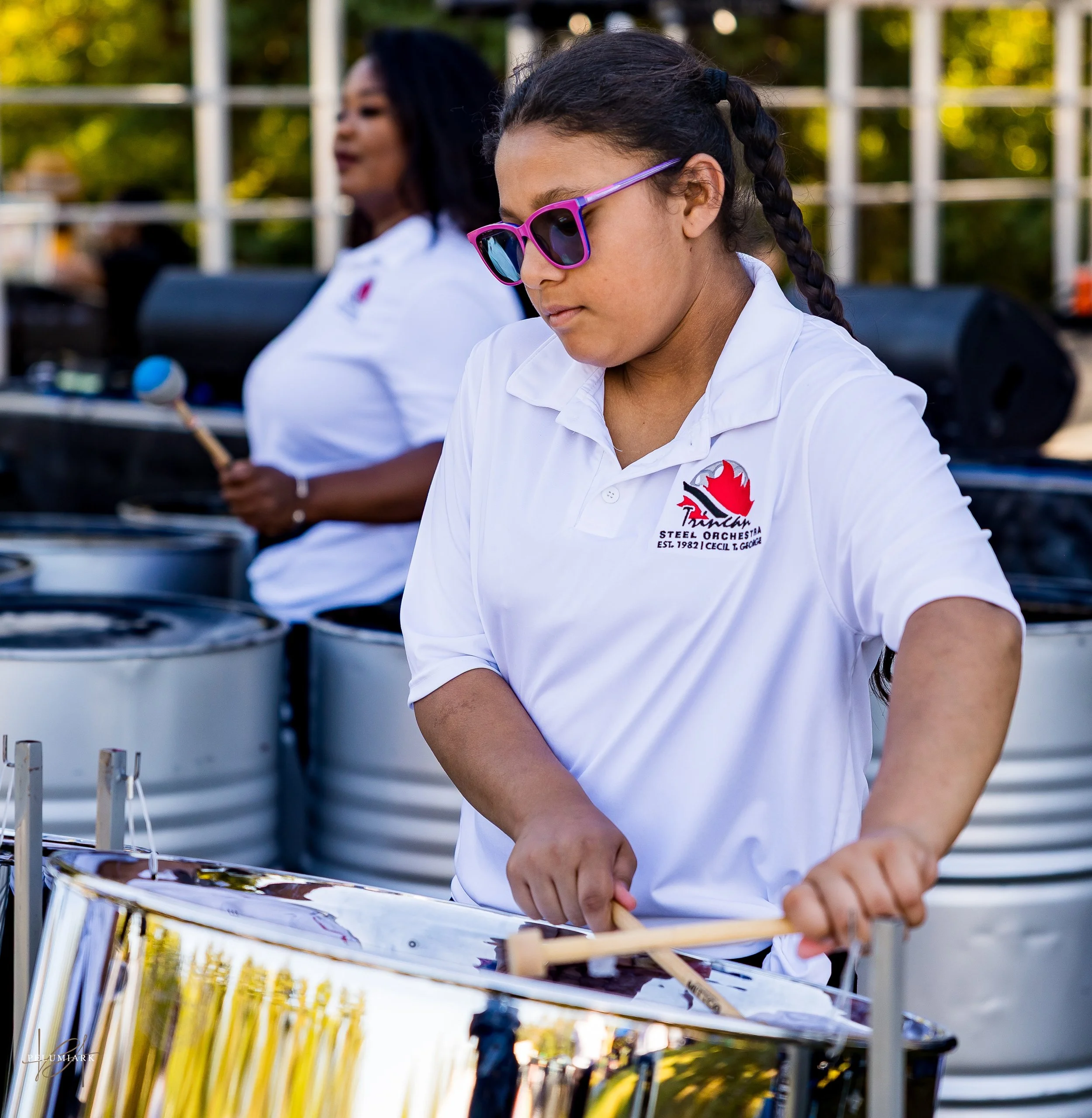 Young girl wearing sunglasses and a white polo shirt playing steel drums outdoors, with another woman in similar attire in the background.