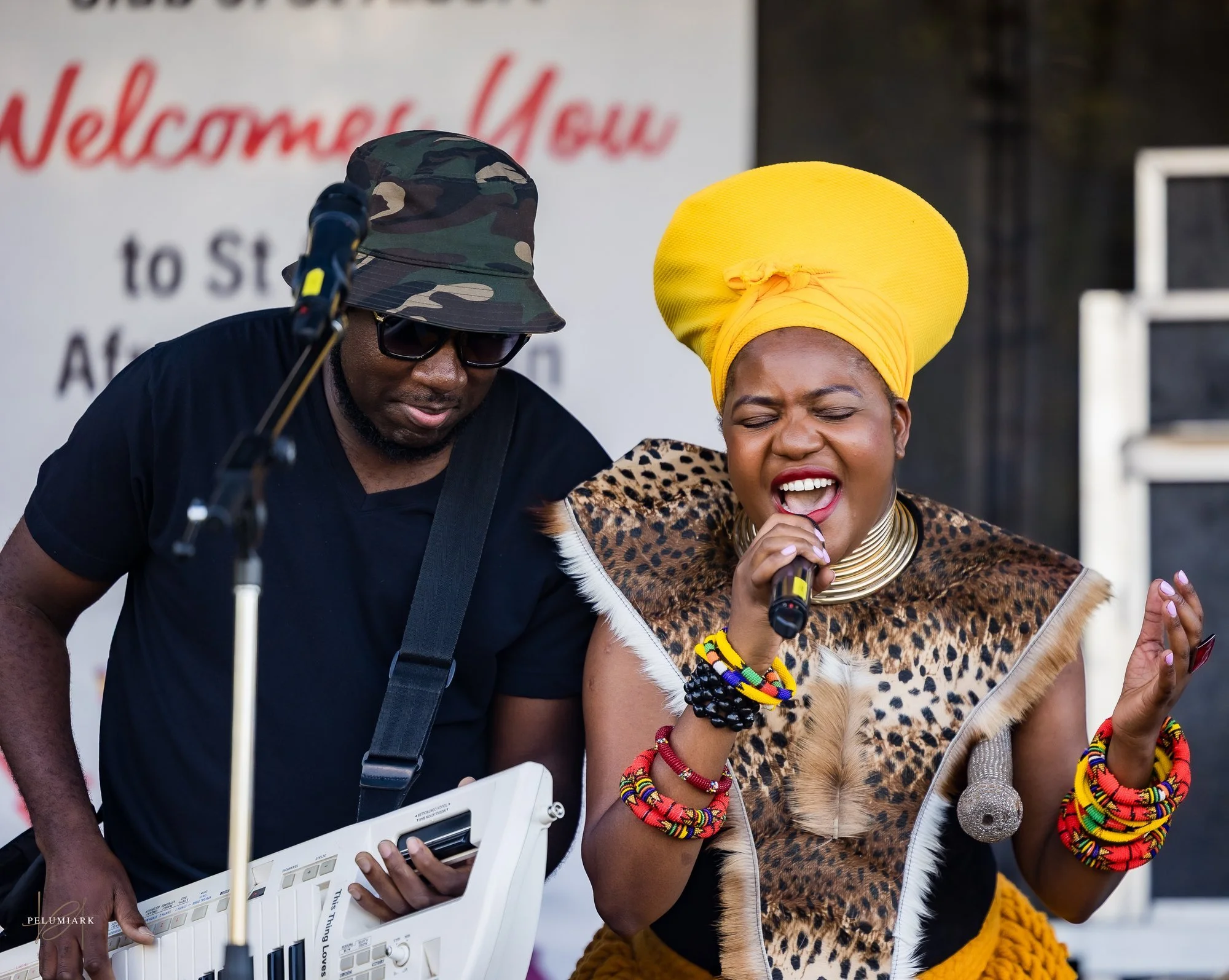 A woman wearing a bright yellow headwrap and colorful jewelry passionately singing into a microphone, accompanied by a man playing an electronic keyboard, both performing on stage.