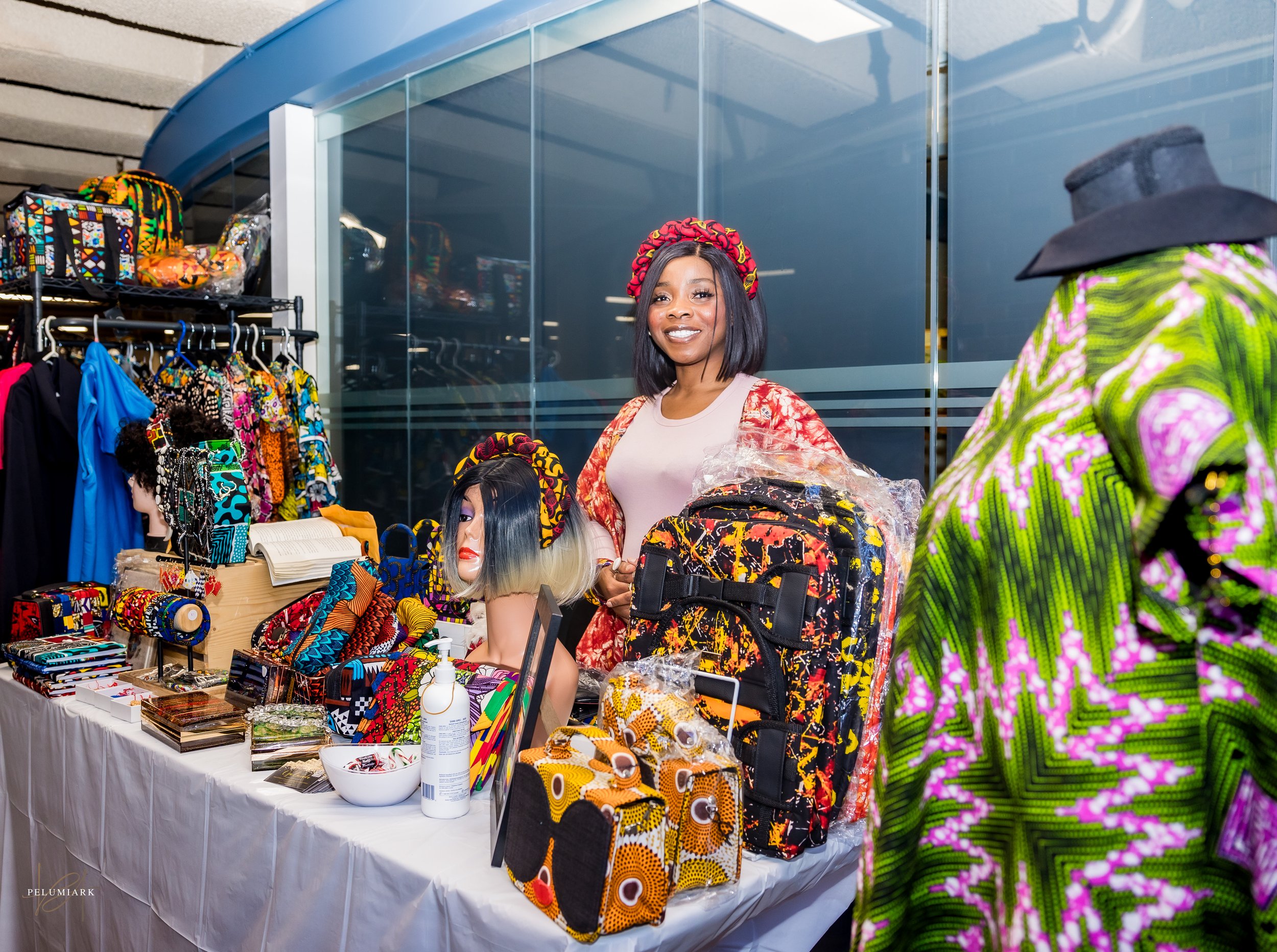 Woman standing behind a table of colorful African-patterned accessories and clothing, at a market stall, with vibrant clothing displayed and mannequins wearing African print garments.