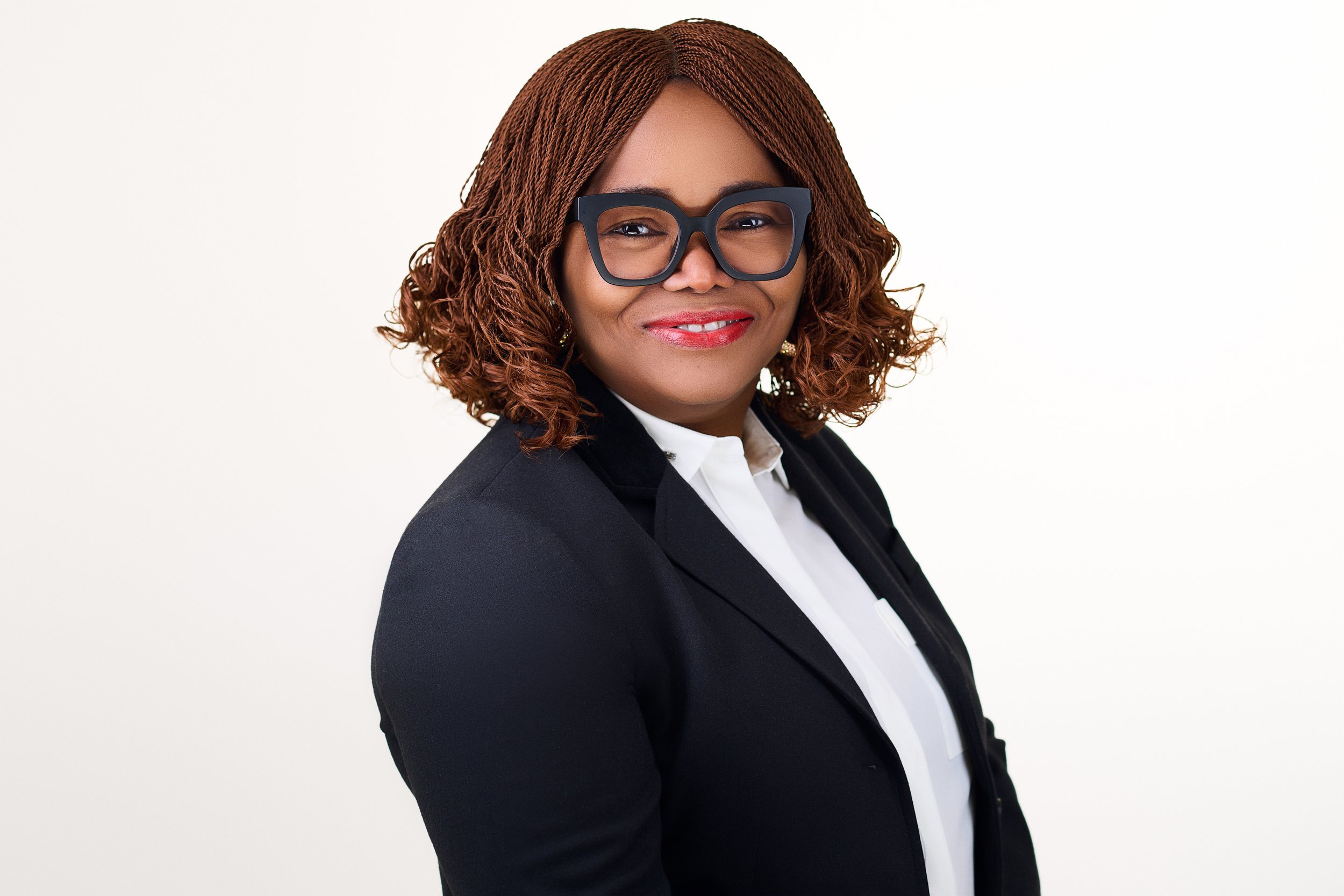 A professional woman with curly, reddish-brown hair, wearing large black glasses, a black blazer, and a white blouse, smiling against a plain white background.